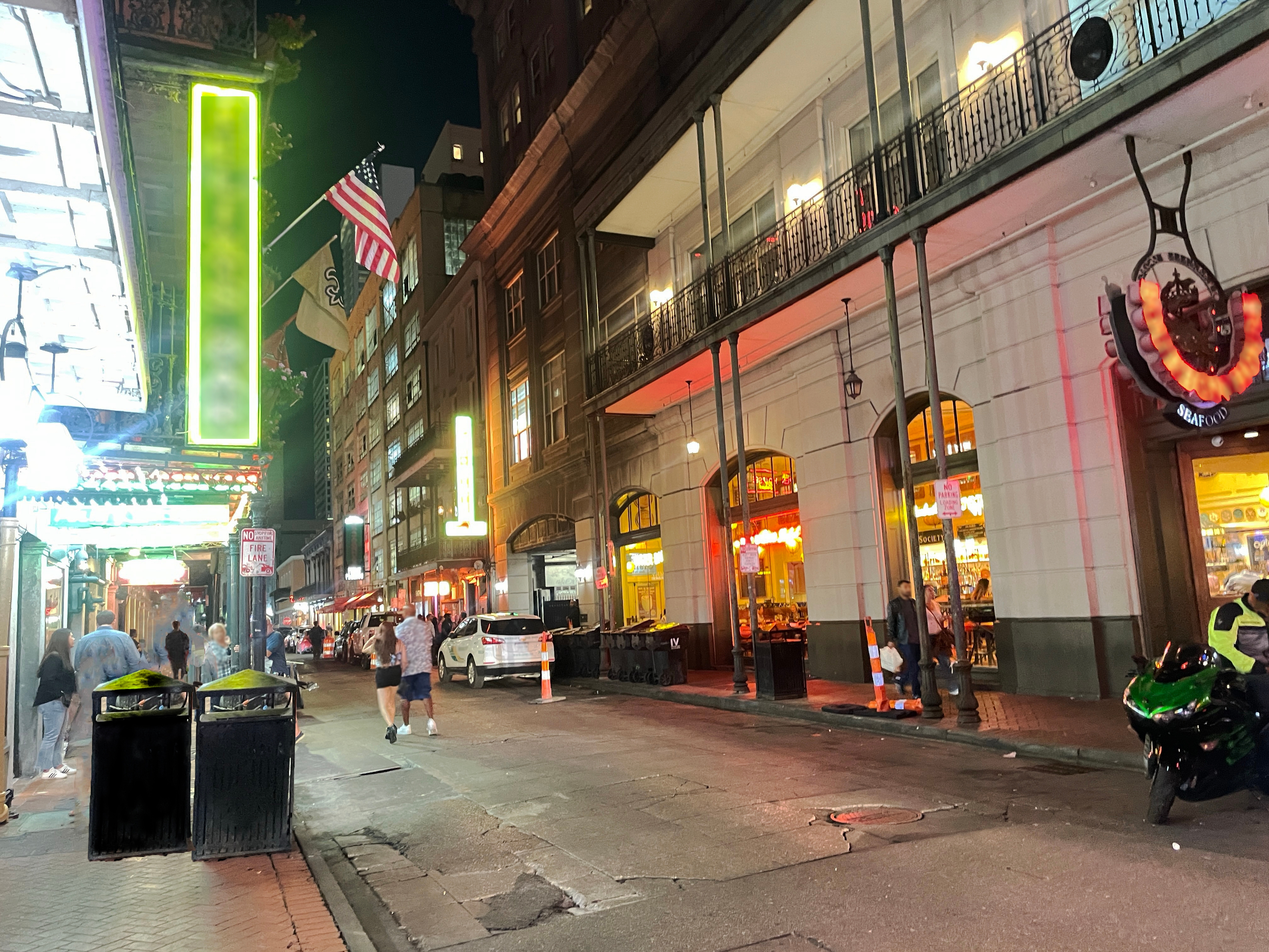 People walking on a lively city street at night with lit storefronts, neon signs, and an American flag