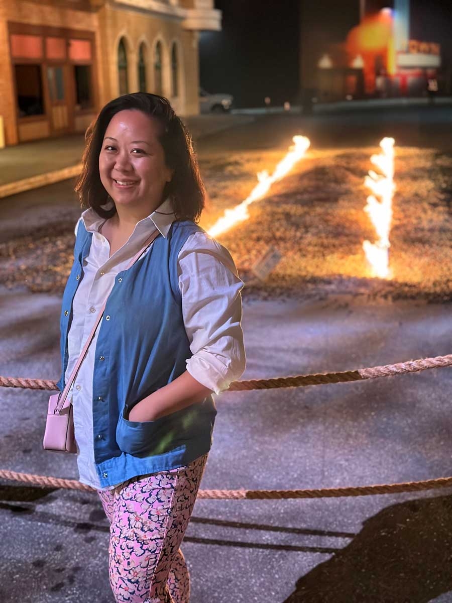 Person smiling outdoors at night, wearing a blue vest over a button-up shirt and patterned pants, standing near flaming lines on the ground