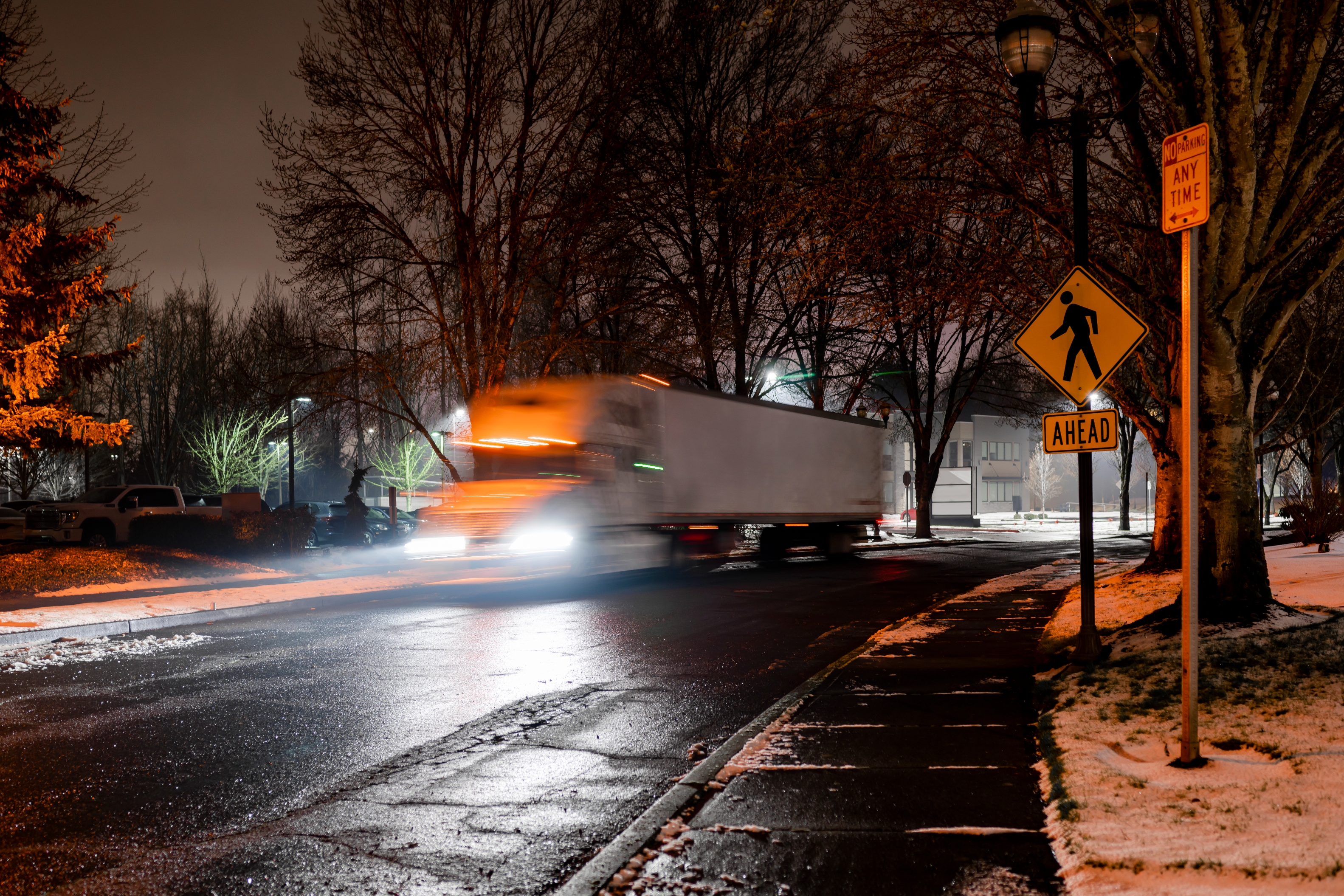 A truck driving on a snowy, dimly lit street with a pedestrian crossing sign visible