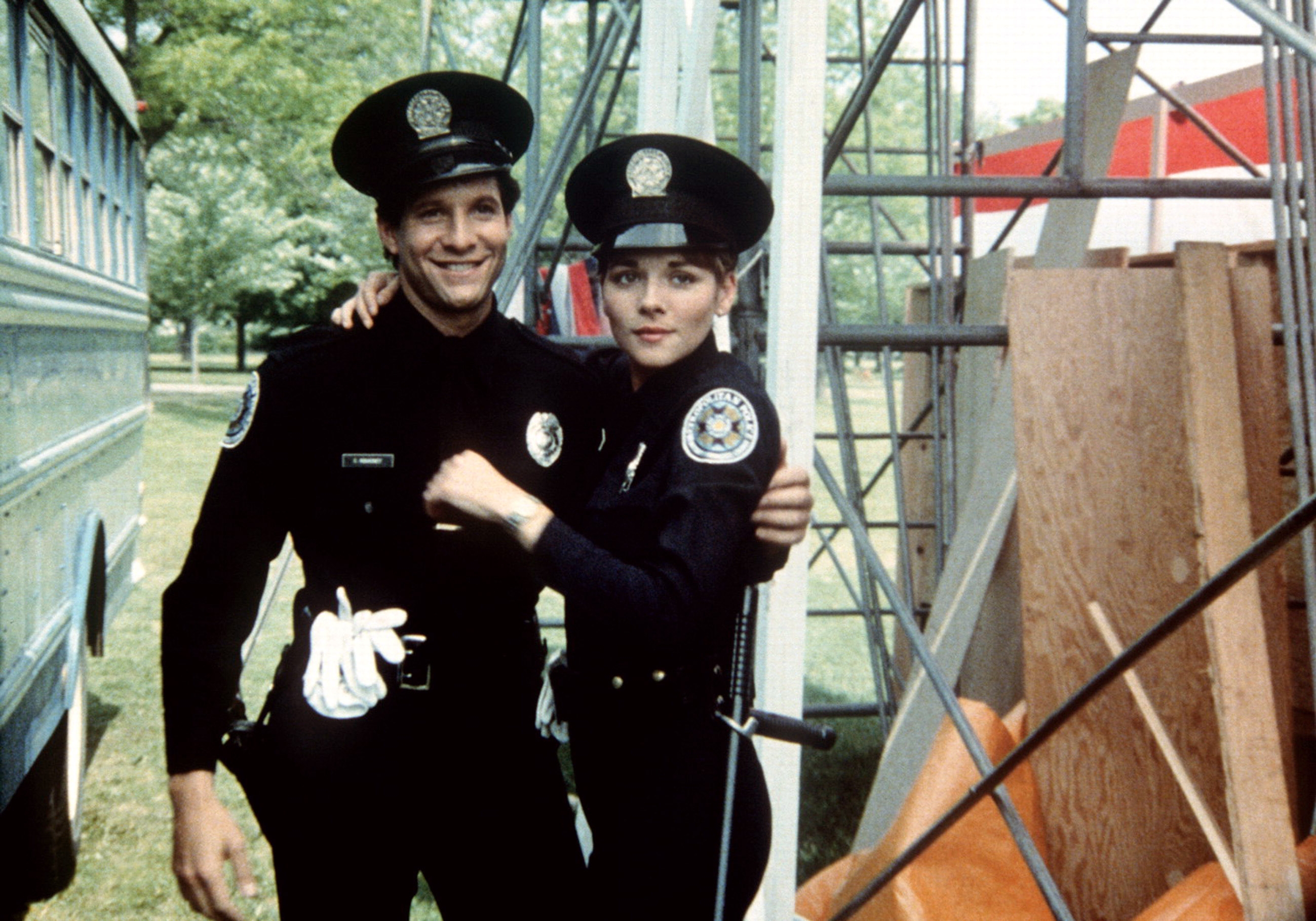 Two actors in police uniforms on a film set, smiling, with scaffolding and equipment in the background