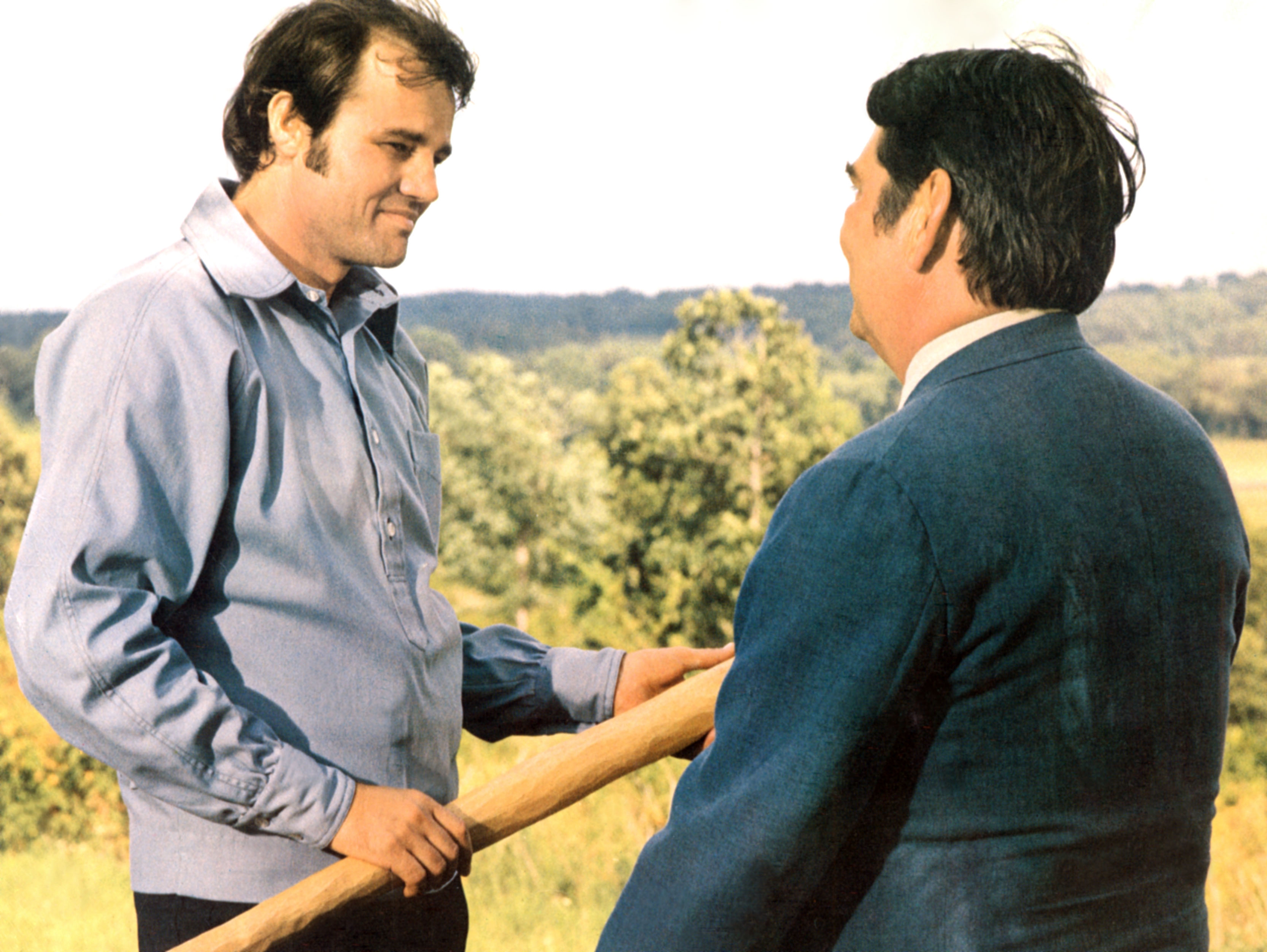 Two men outdoors amicably holding a baseball bat, one in a casual shirt and the other in a suit, engaging in conversation