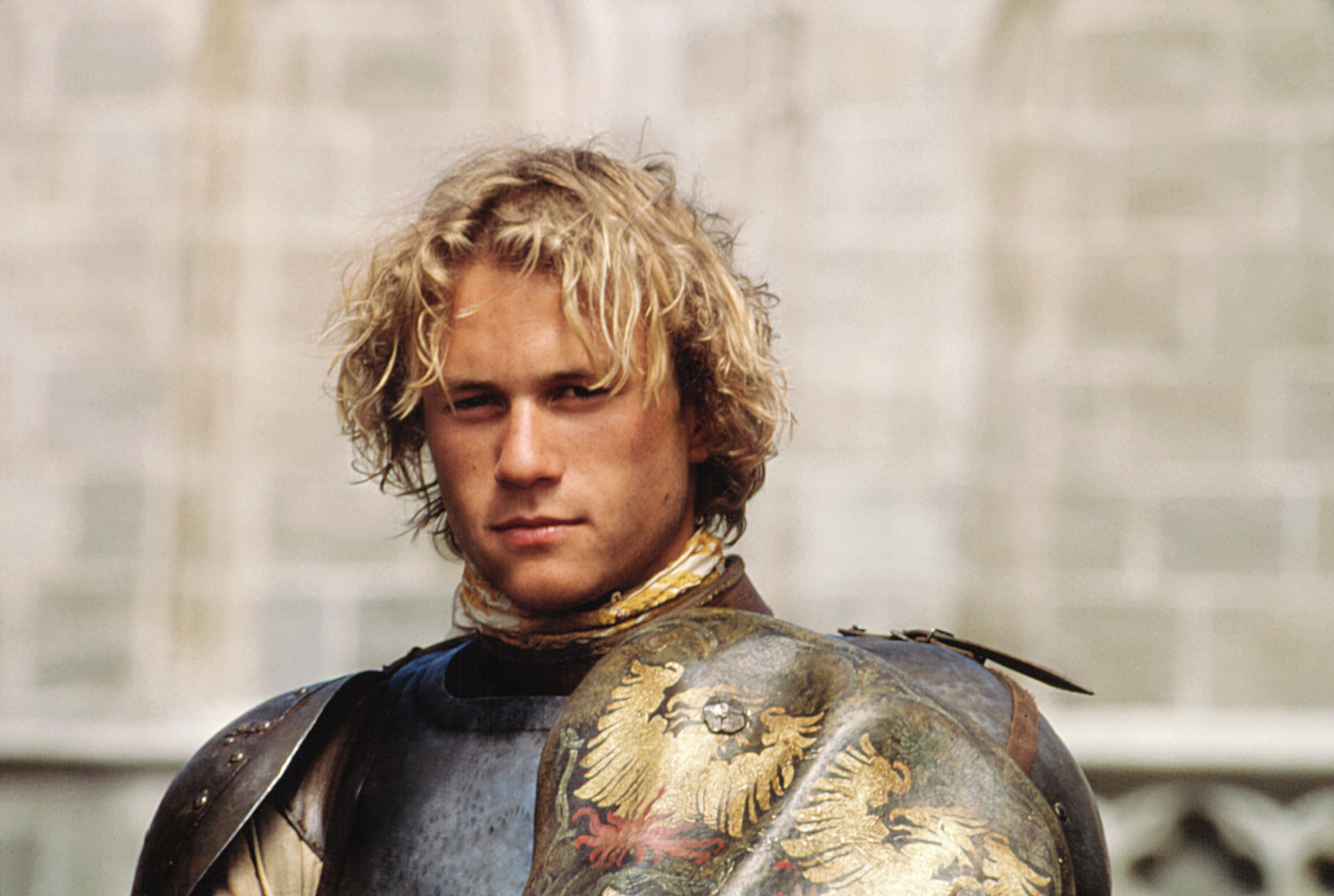 Person in medieval armor with tousled hair, standing against a stone wall backdrop. The scene evokes a historical or fantasy setting