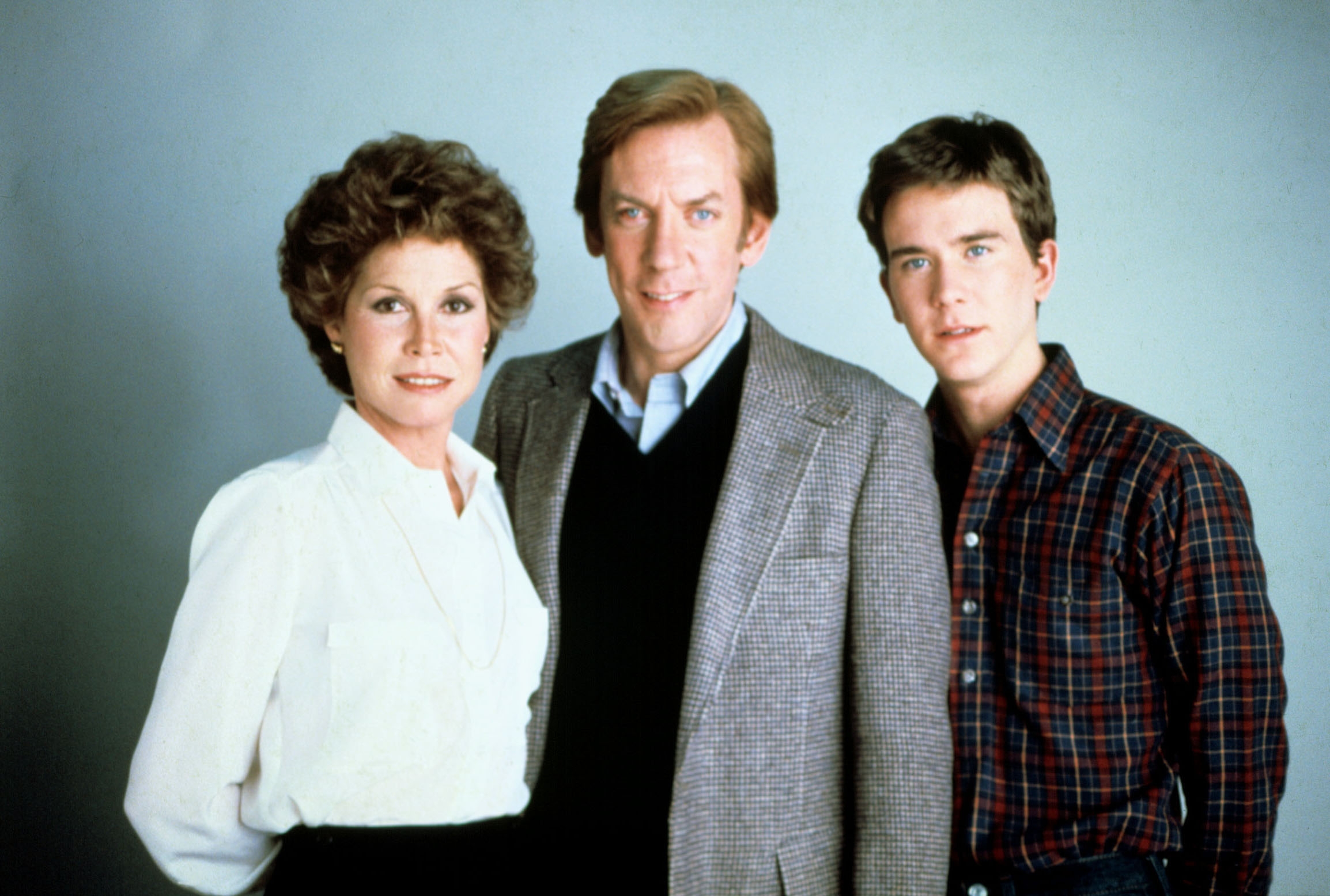 Three people posed together; one in a white blouse, one in a blazer and sweater, and one in a plaid shirt, conveying a classic TV family vibe