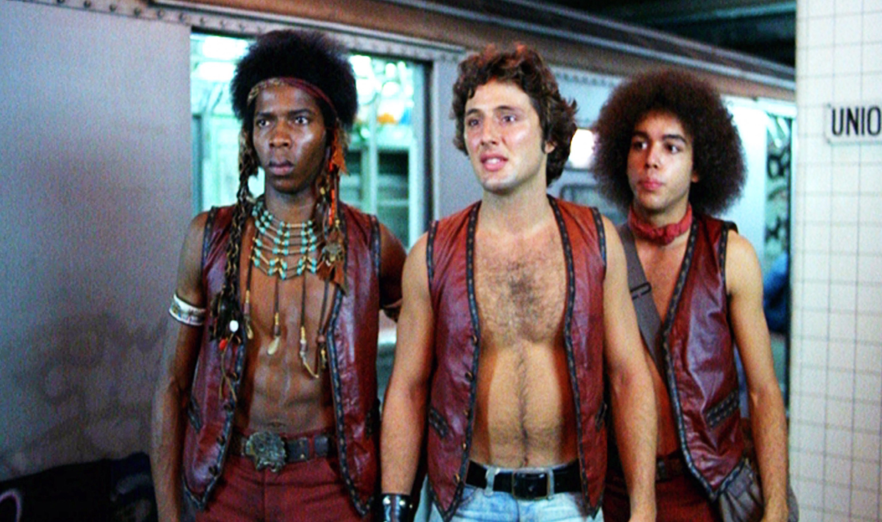 Three men in a subway station wearing vintage-style leather vests and accessories, evocative of a 1970s film scene