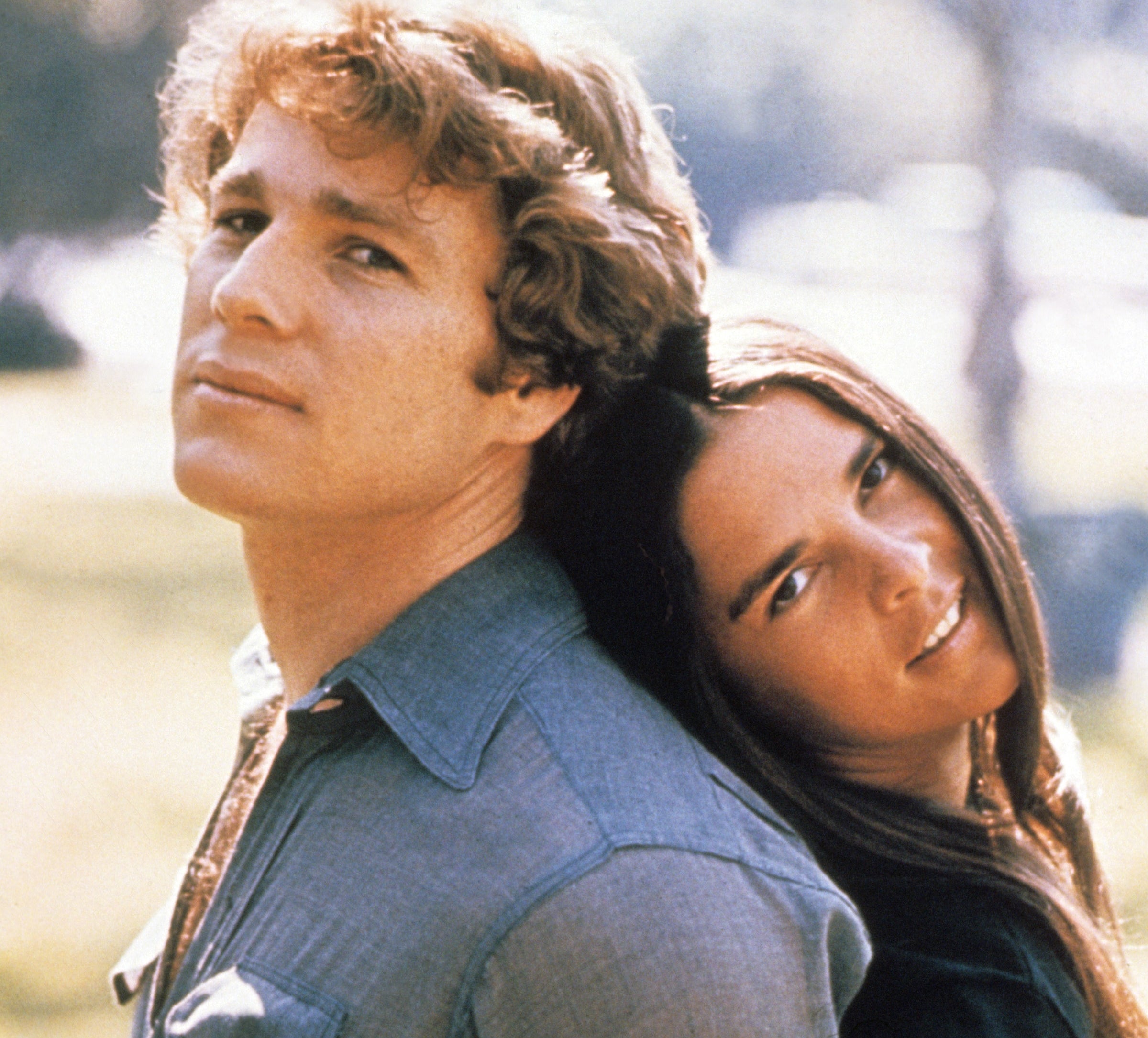 A man and woman stand back-to-back, outdoors. The woman leans her head on his shoulder, both with relaxed expressions, creating a nostalgic vibe