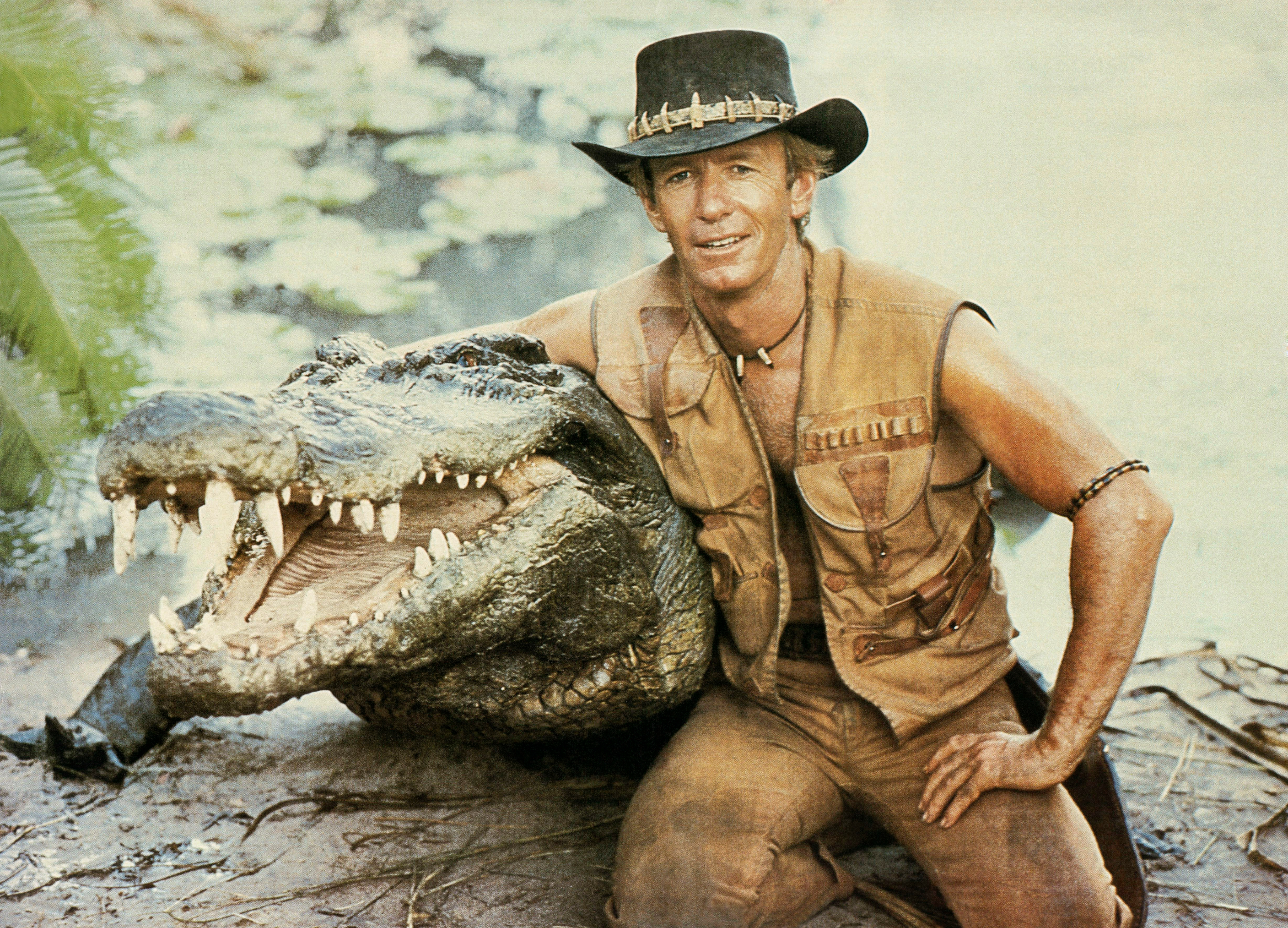 Man in outdoor attire and hat poses with large crocodile in a natural setting, promoting adventurous TV/movie theme