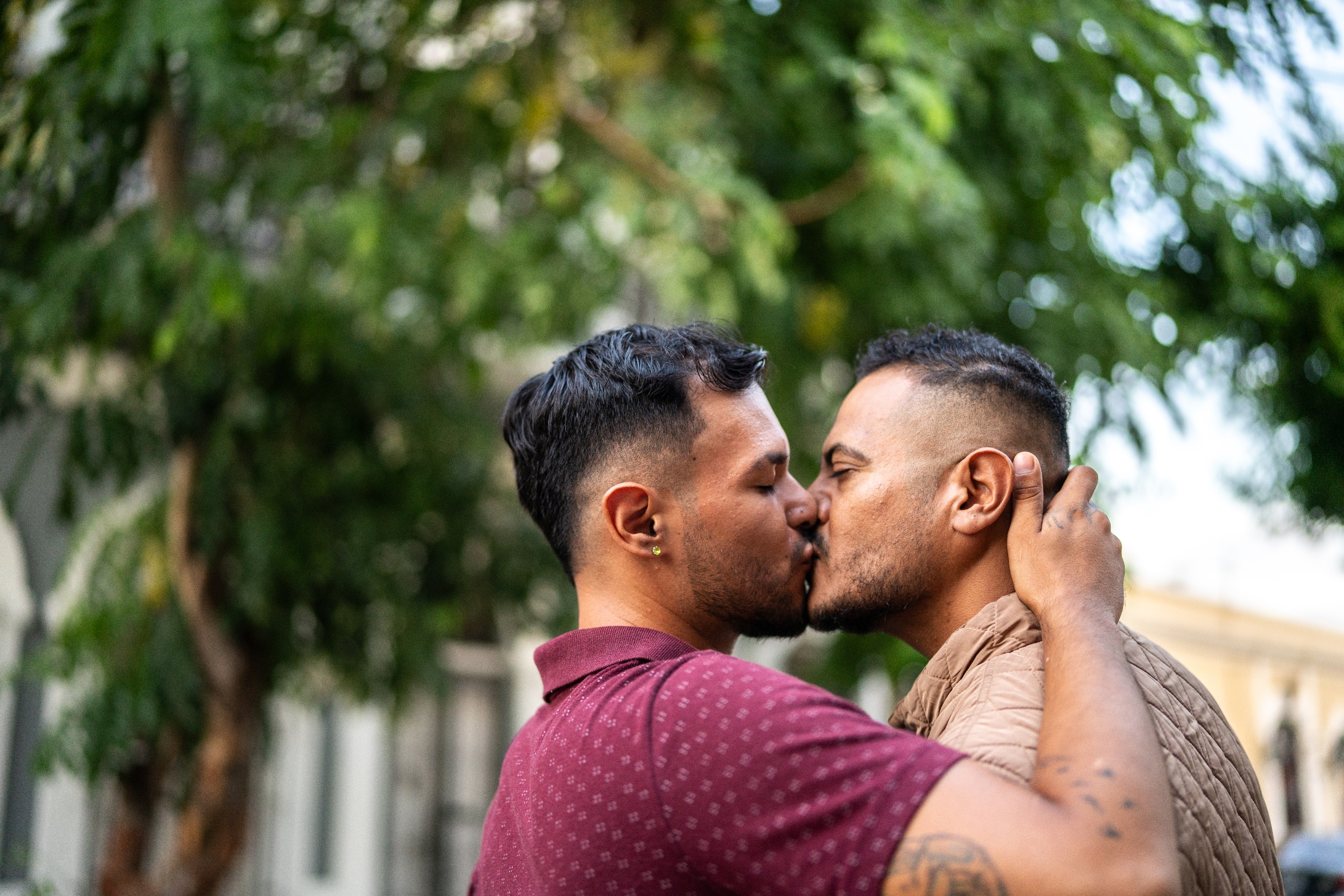 Two people sharing a romantic kiss outdoors, with greenery in the background. One wearing a patterned shirt, the other a textured jacket