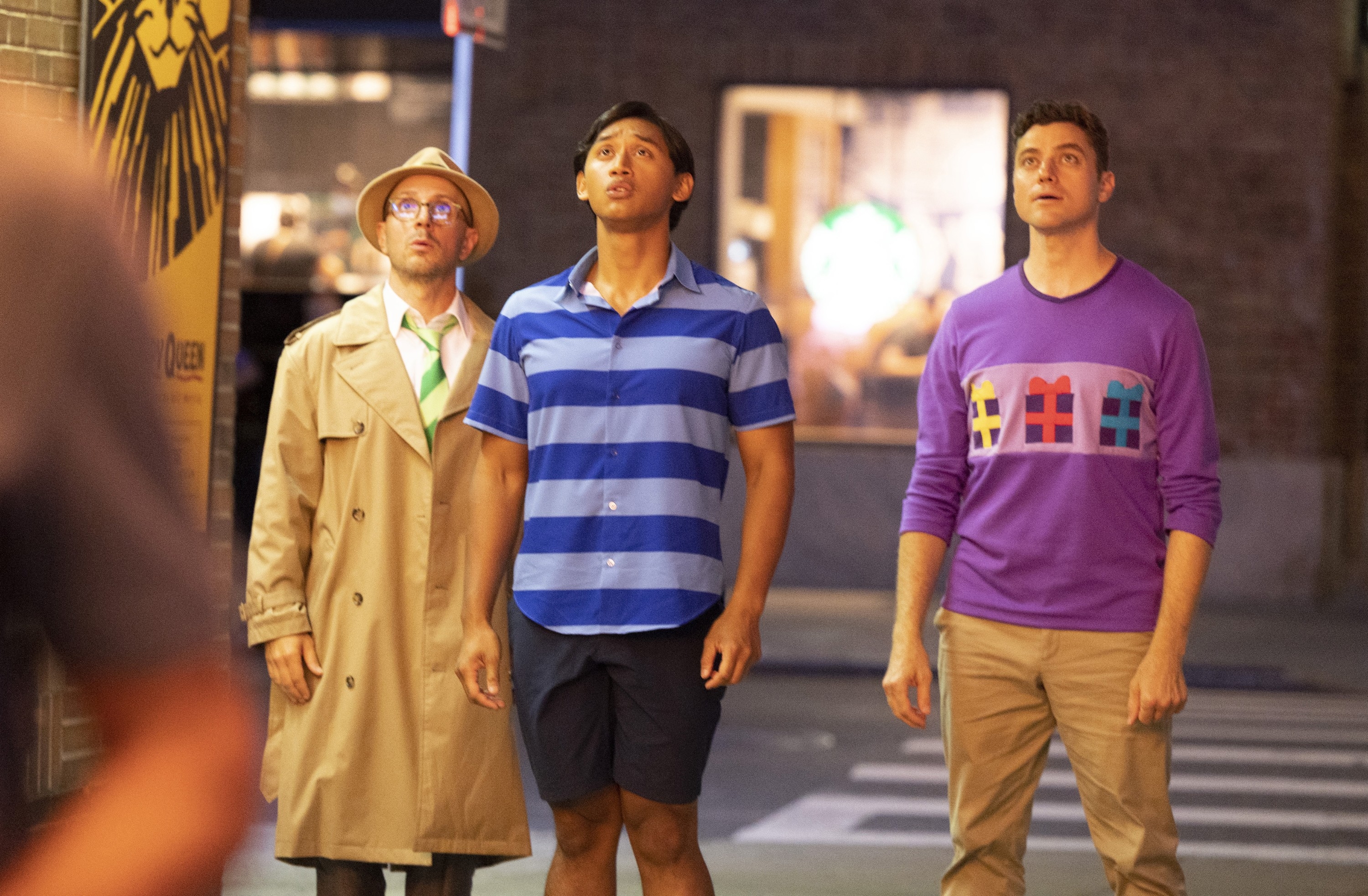 Three people on a city street look upwards. One wears a trench coat, another a striped polo, and the third a graphic sweater