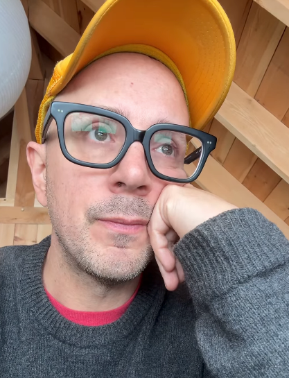 Person wearing glasses and a cap, gazing thoughtfully, with chin resting on hand in a cozy wooden interior