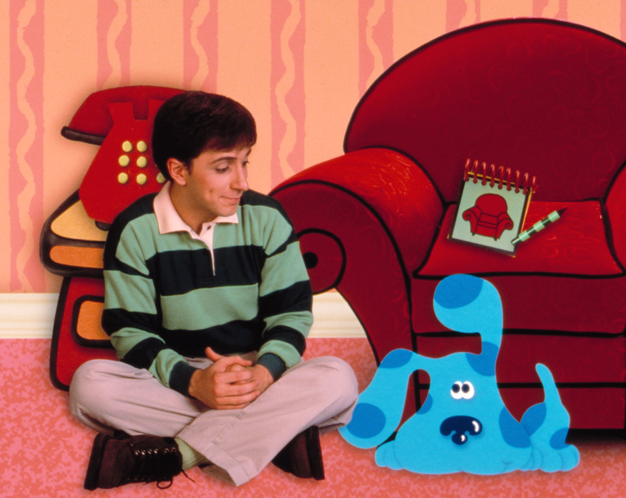 Person in a striped shirt sits cross-legged on the floor next to a cartoon blue dog, in a cozy room with a red couch and a toy phone