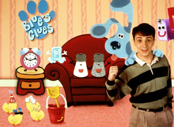 A young man in a striped shirt stands with animated characters from a children's TV show, including a blue dog and various household items with faces