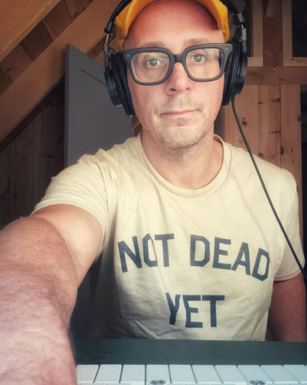 Person wearing glasses and headphones, in a room with wood paneling, wearing a t-shirt with "Not Dead Yet" printed on it