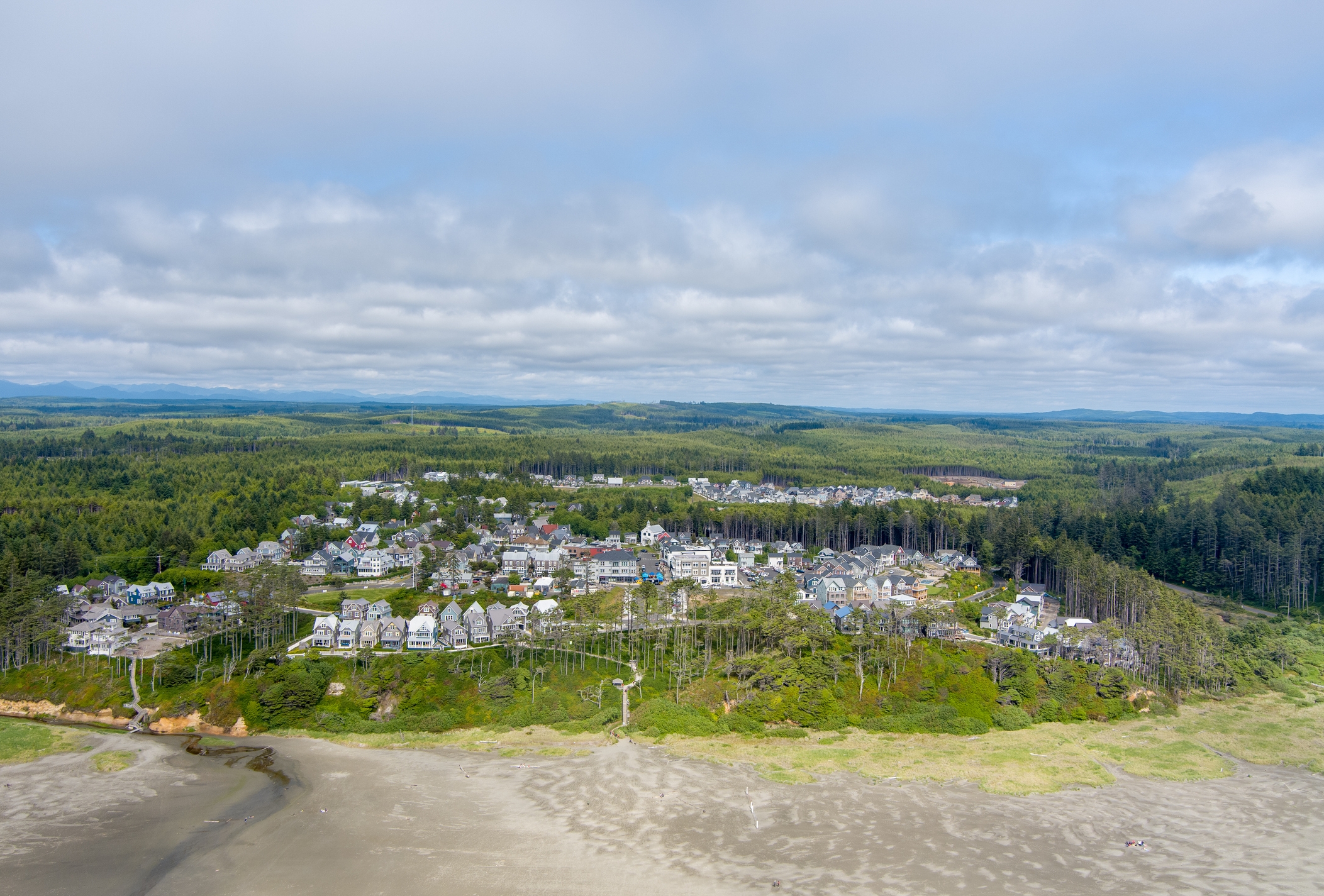 Aerial view of a coastal town surrounded by greenery, with houses near the beach and a forest in the background under a cloudy sky