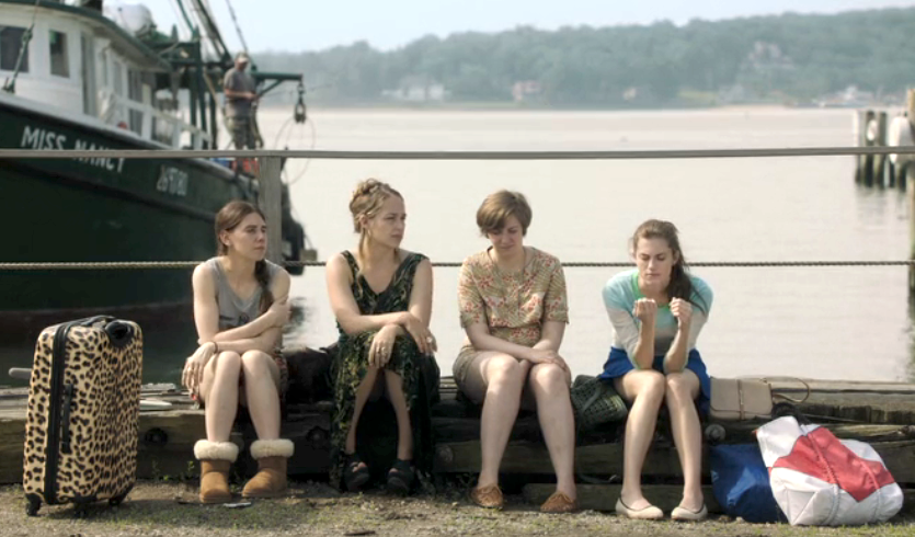 four girls sit on a dock with a suitcase A boat is in the background