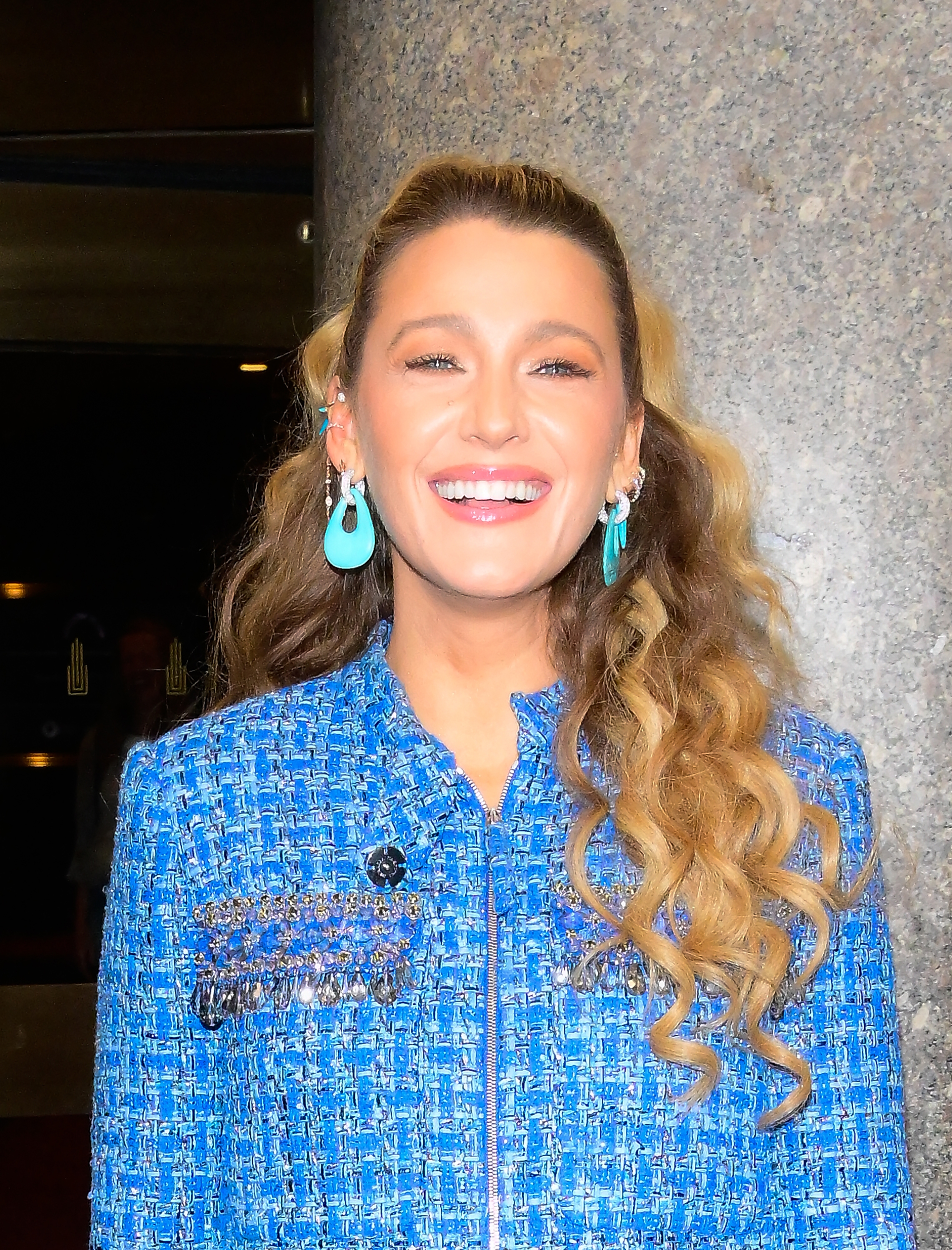 Blake Lively in a textured outfit smiles, wearing turquoise earrings, against a stone wall background