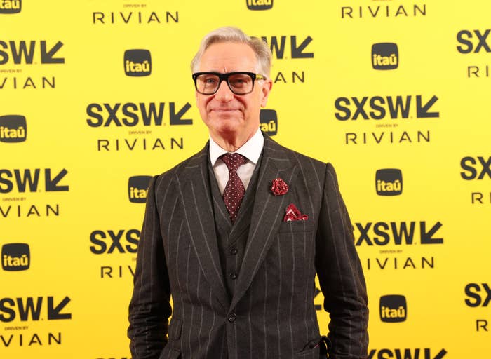 Paul Feig in pinstripe suit and tie, with glasses and pocket square, attends SXSW event. Yellow backdrop with event logos