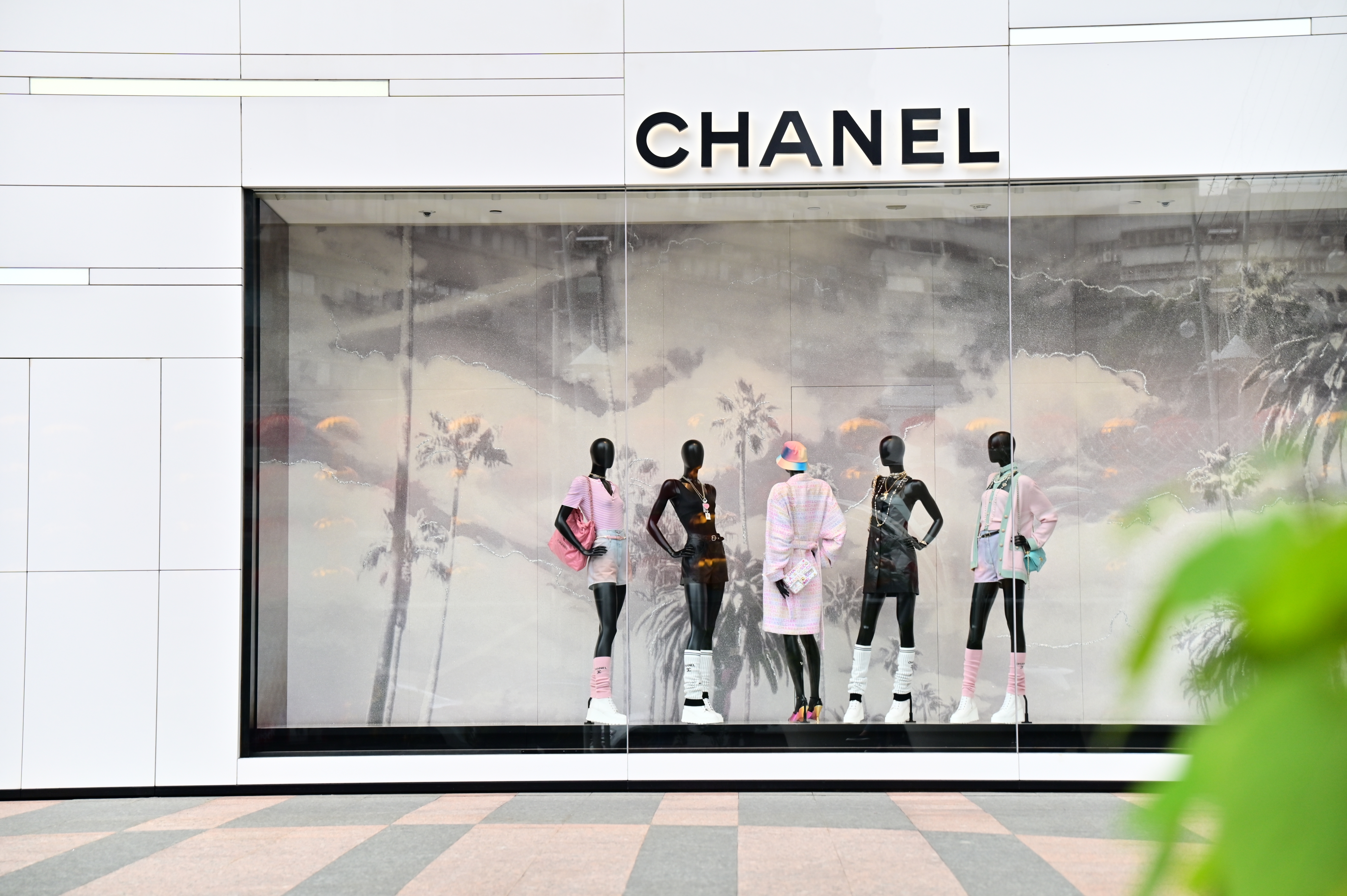 Five mannequins in stylish outfits are displayed in a Chanel store window with a tropical backdrop