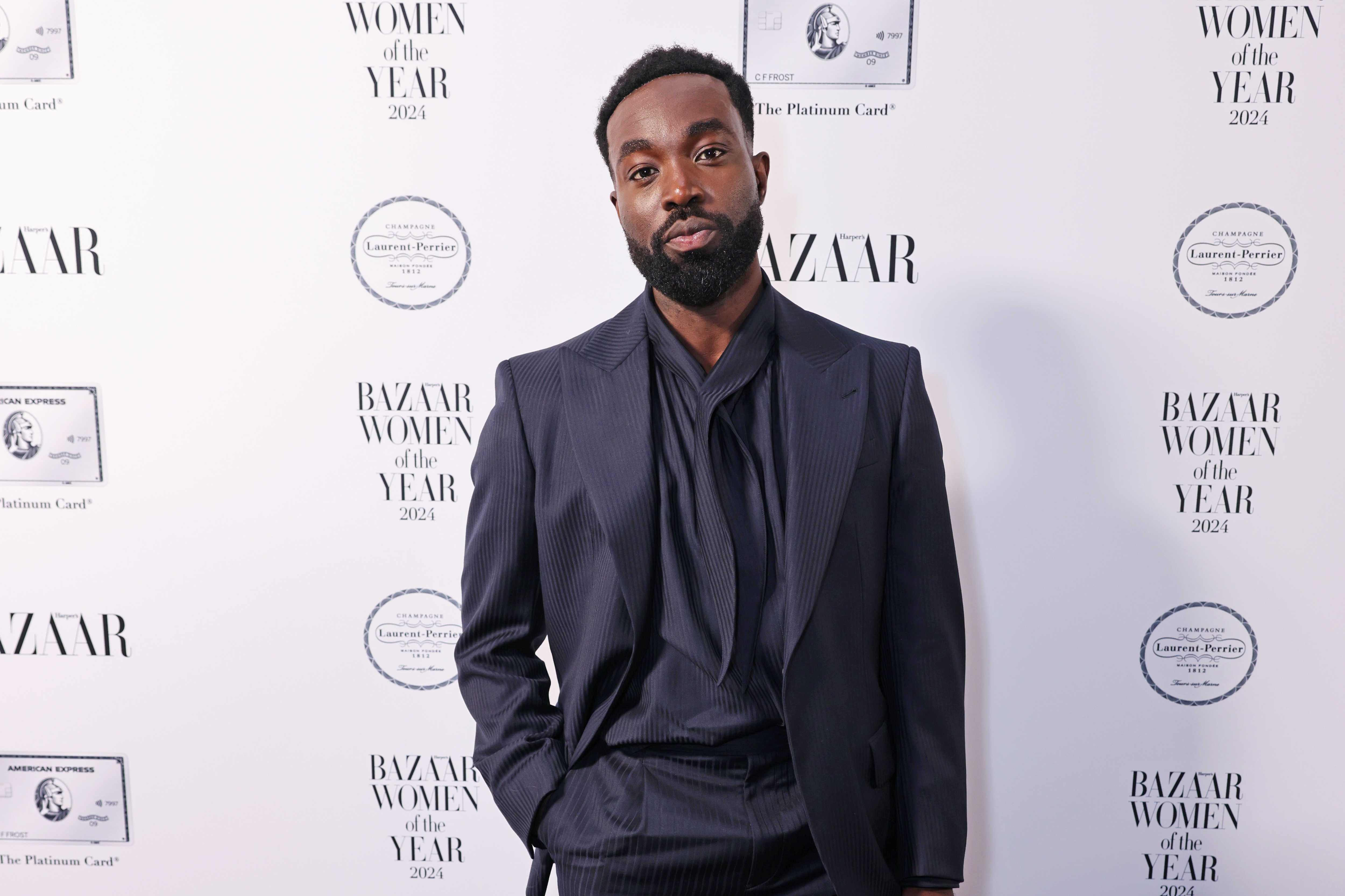 Paapa Essiedu in a tailored dark suit stands in front of a &quot;Bazaar Women of the Year 2023&quot; event backdrop