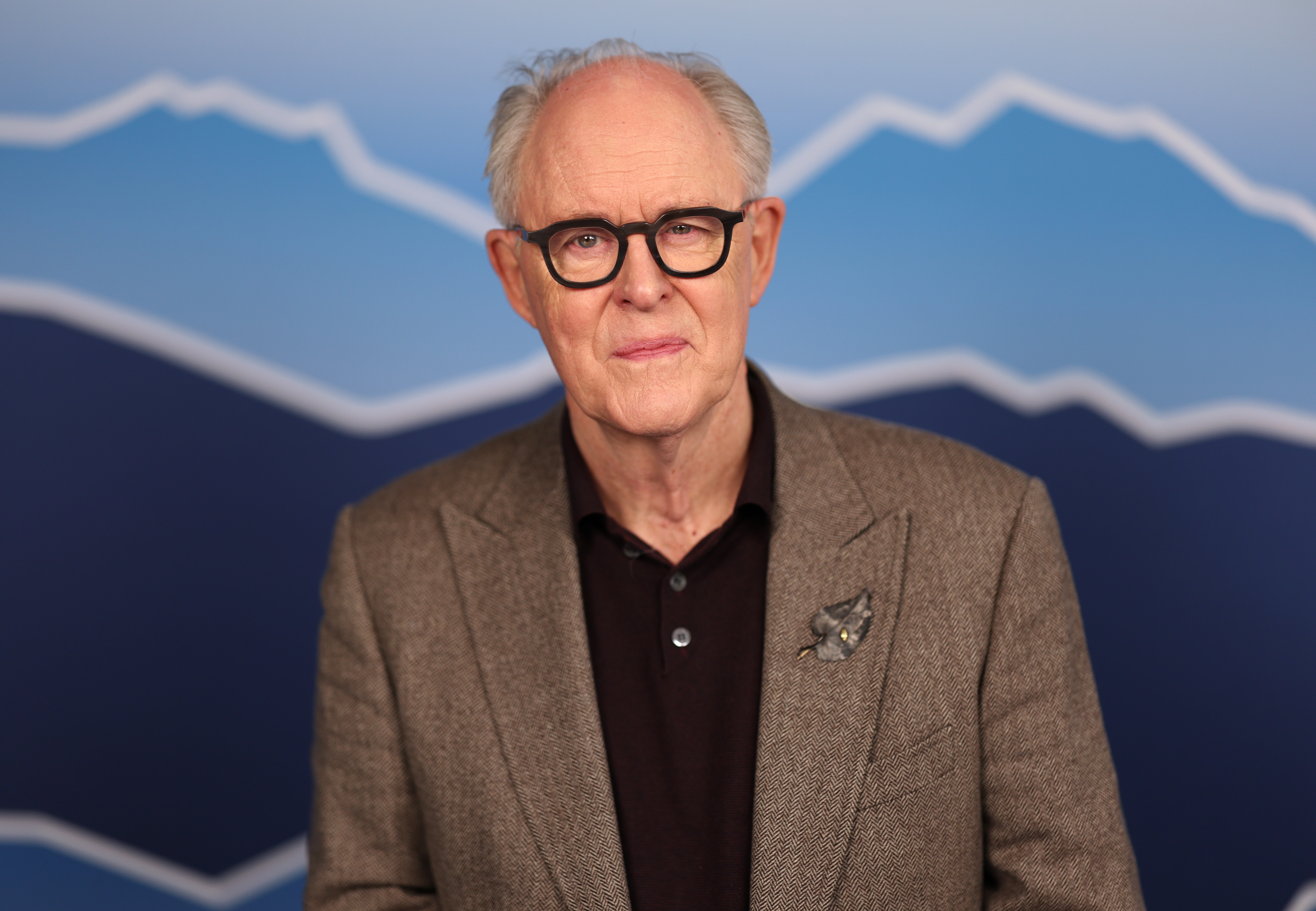 John Lithgow in a tweed blazer with a star-shaped pin, wearing glasses, stands in front of a mountain-themed backdrop