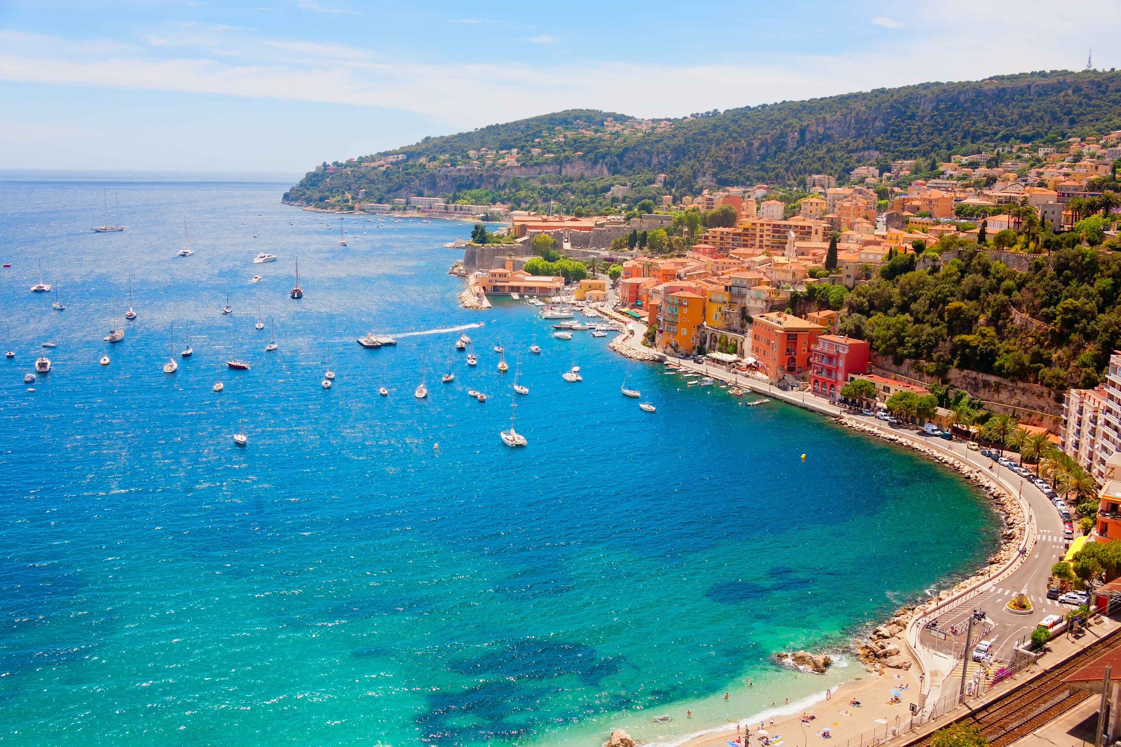 Coastal town with hillside buildings, a curved shoreline, and sailboats on the blue sea under a clear sky