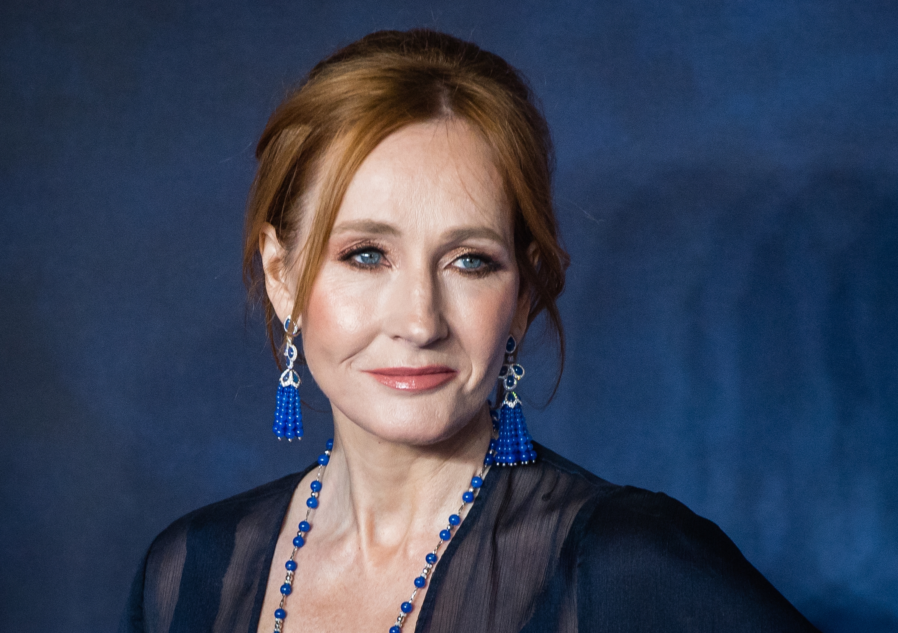J.K. Rowling with long hair and blue beaded jewelry, wearing a sheer top, looking towards the camera