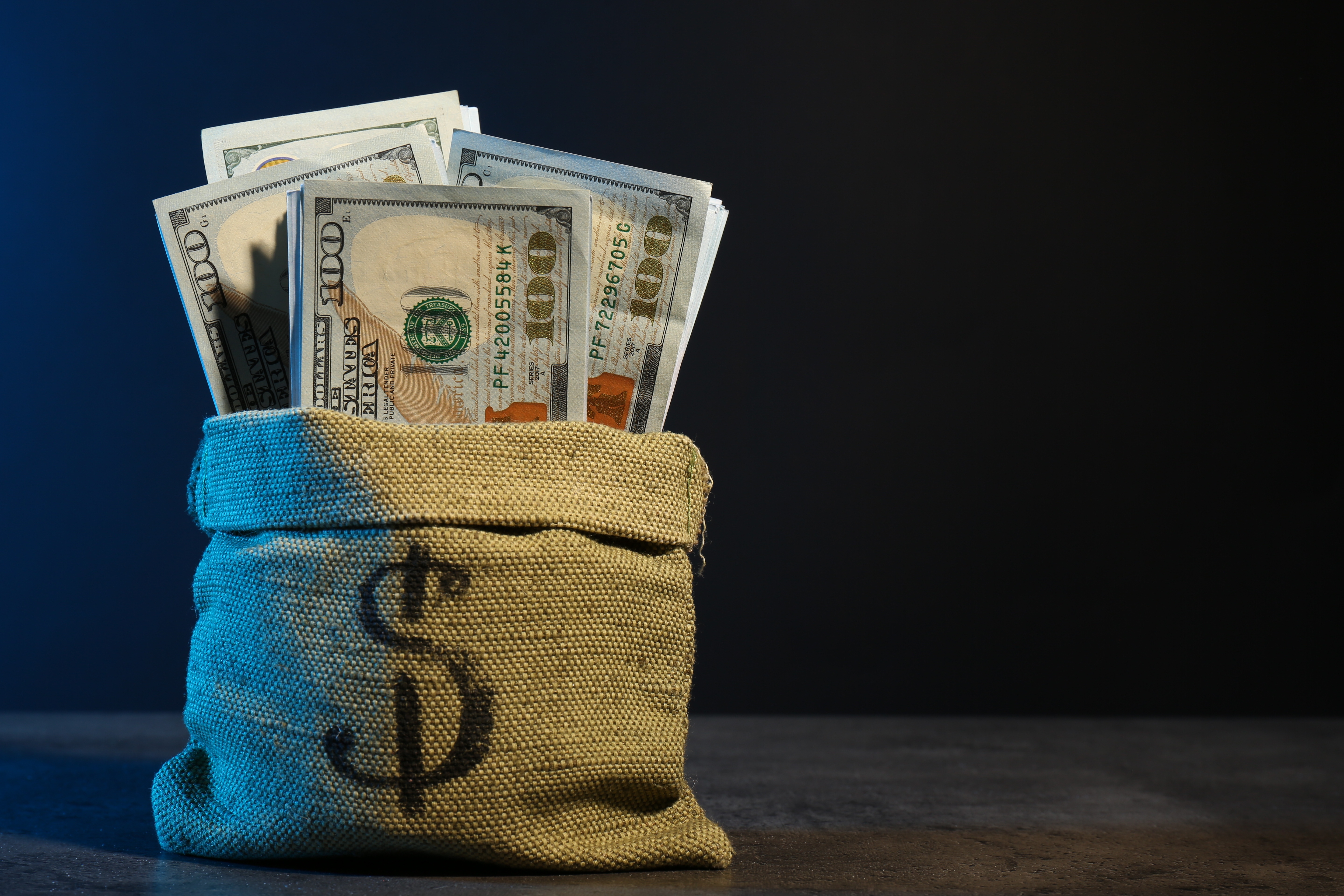 A burlap sack with a dollar sign is filled with U.S. banknotes, standing on a table against a plain background