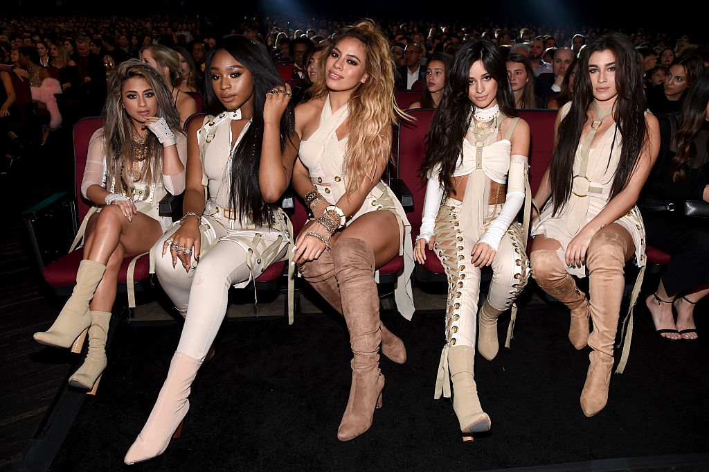 Five women in coordinated, stylish outfits are seated at an event, likely a performance or awards show