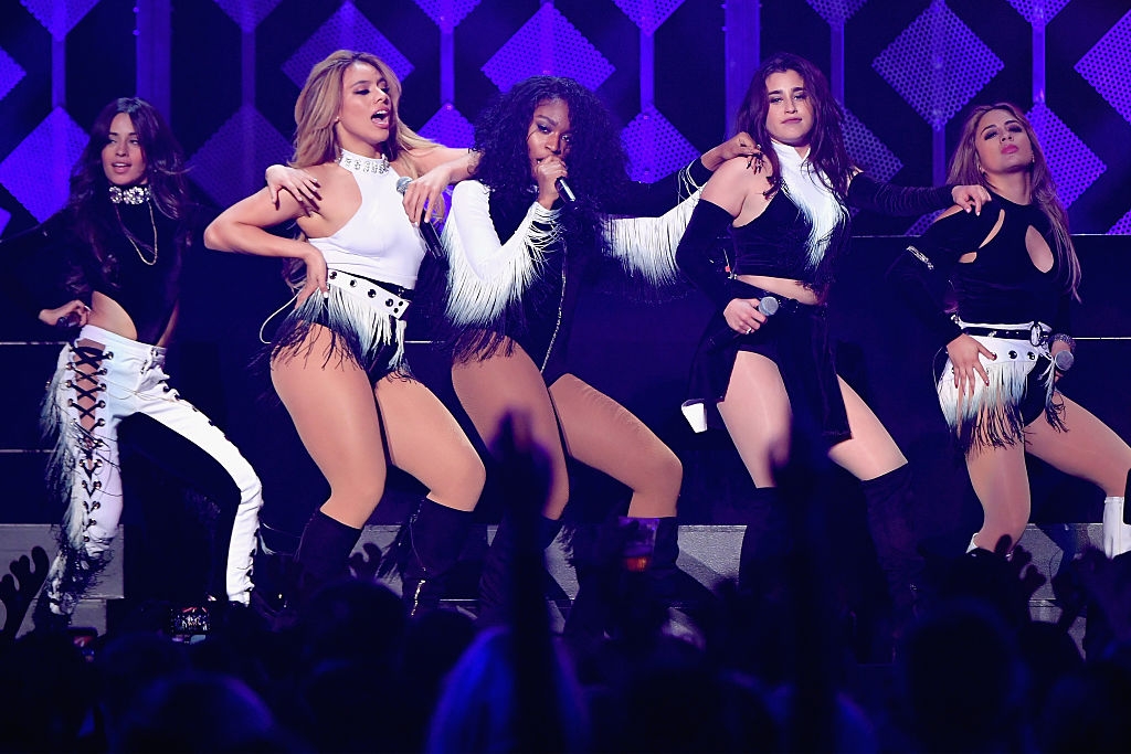 Group of five performers on stage, wearing coordinated black and white outfits with fringe details, dancing energetically in front of a crowd