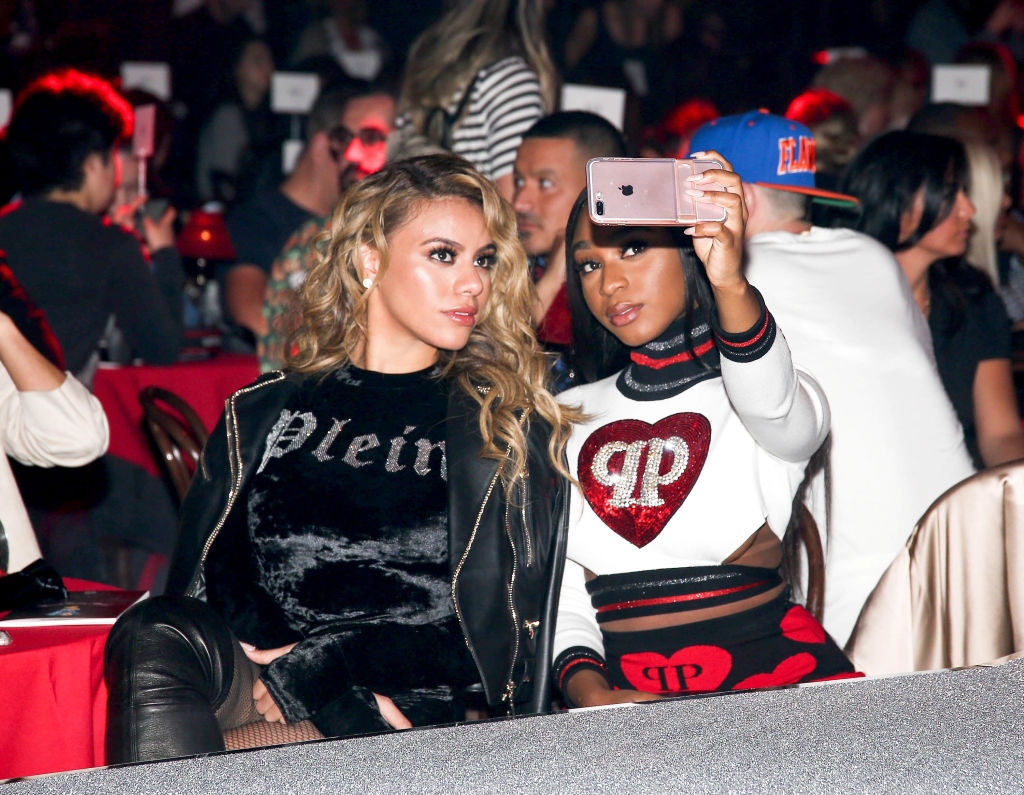 Two women seated at an event. One woman in a velvet top gazes ahead, while the other in a heart-themed outfit takes a selfie with her phone