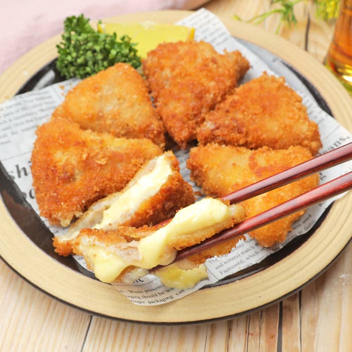 チーズが溶けたフライドコロッケが円形に並んだ皿に盛られ、割った一つが箸で持たれている。皿にはパセリとレモンが添えられている。