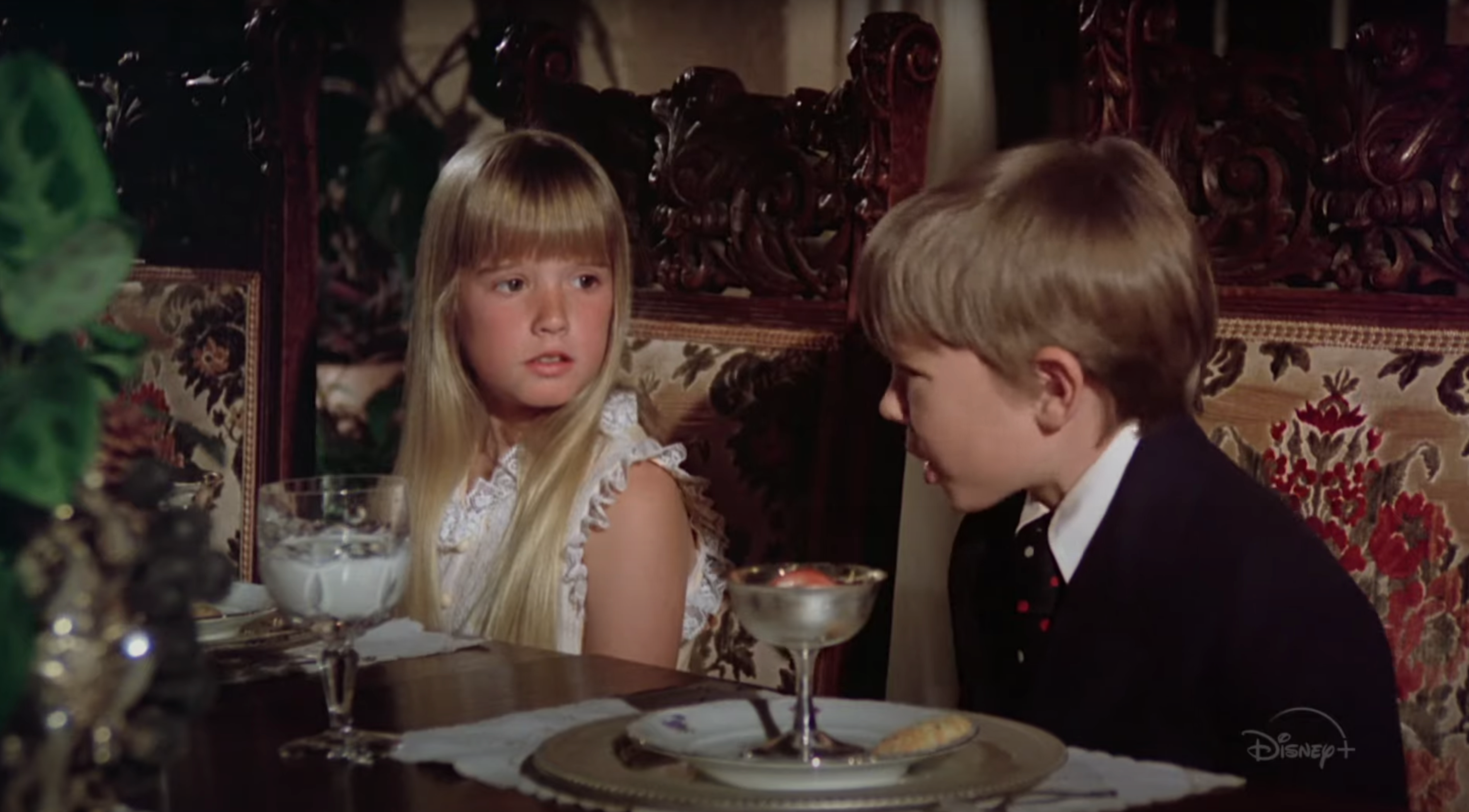 Two children at a fancy dining table, a girl with long hair and a boy in a suit, in conversation