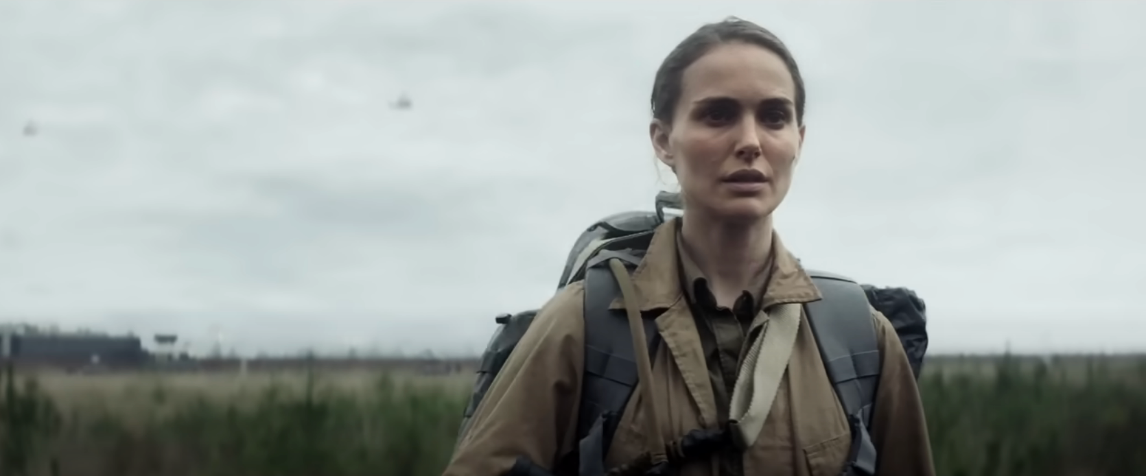 In a scene from "Annihilation," Natalie Portman walks through a field against a clouded sky with hovering aircraft