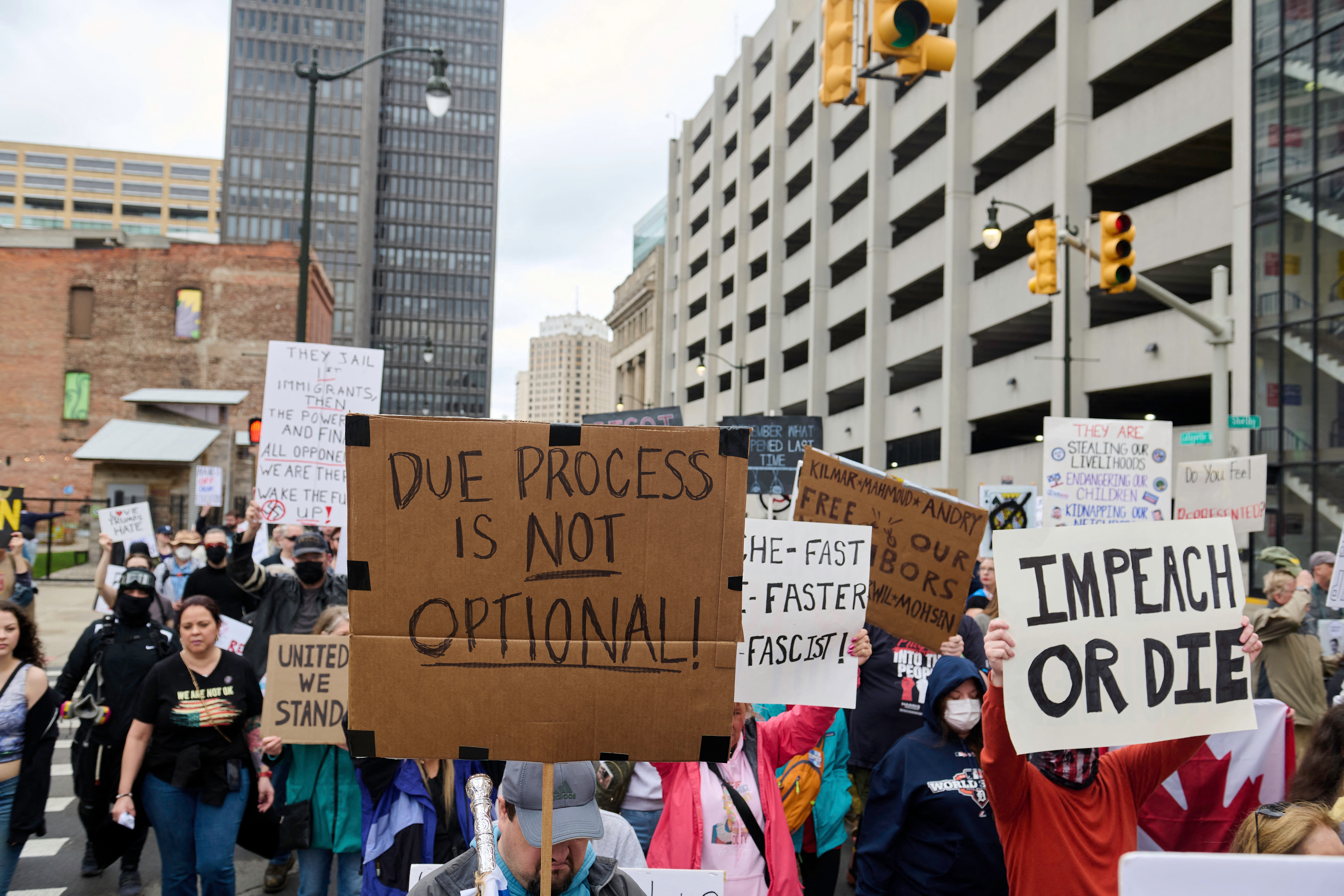 People at a protest hold signs with messages like "Due Process is Not Optional" and "Impeach or Die" in an urban setting