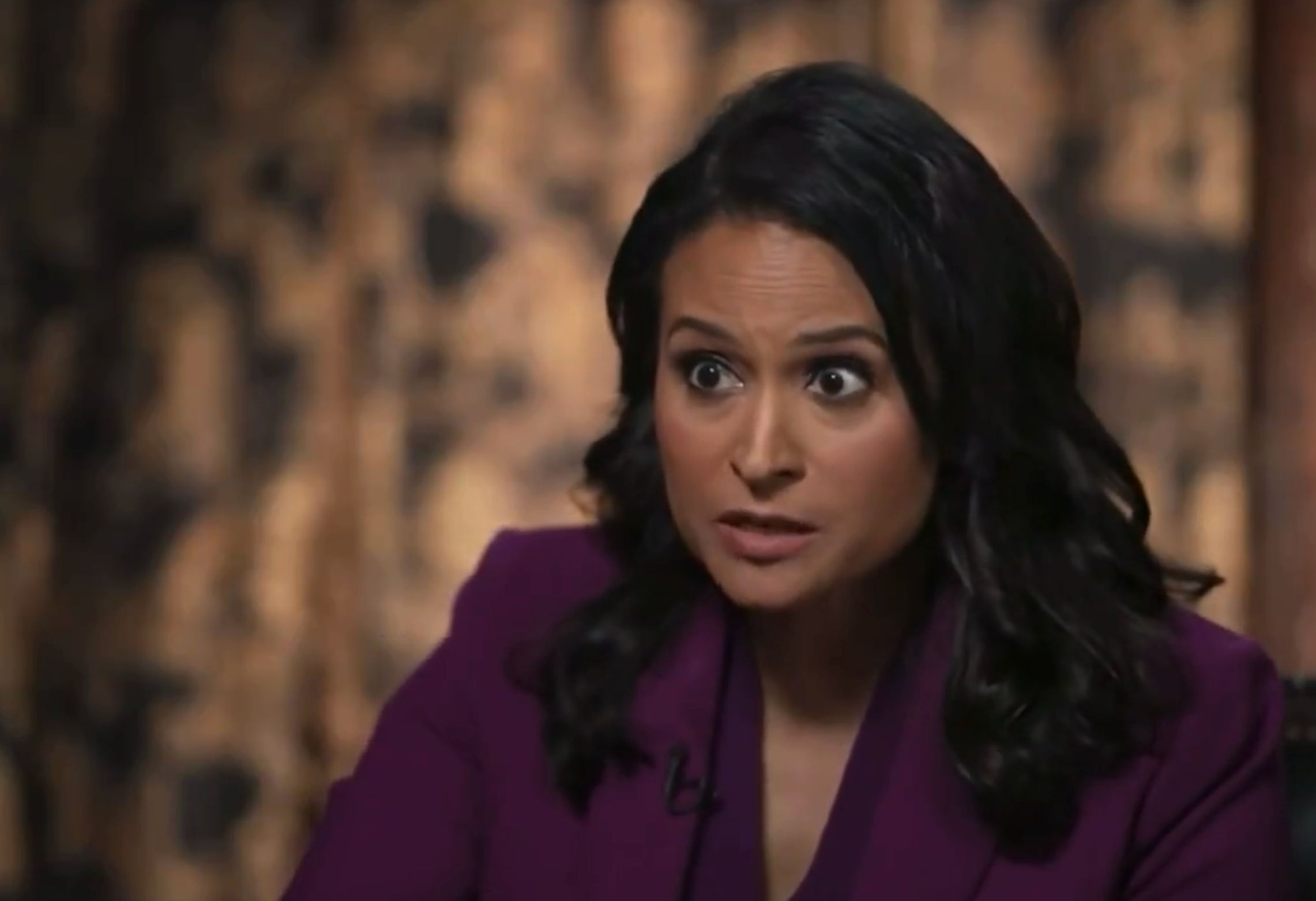 A person in a professional setting looks engaged in conversation, wearing a tailored suit. Their expression is serious and attentive