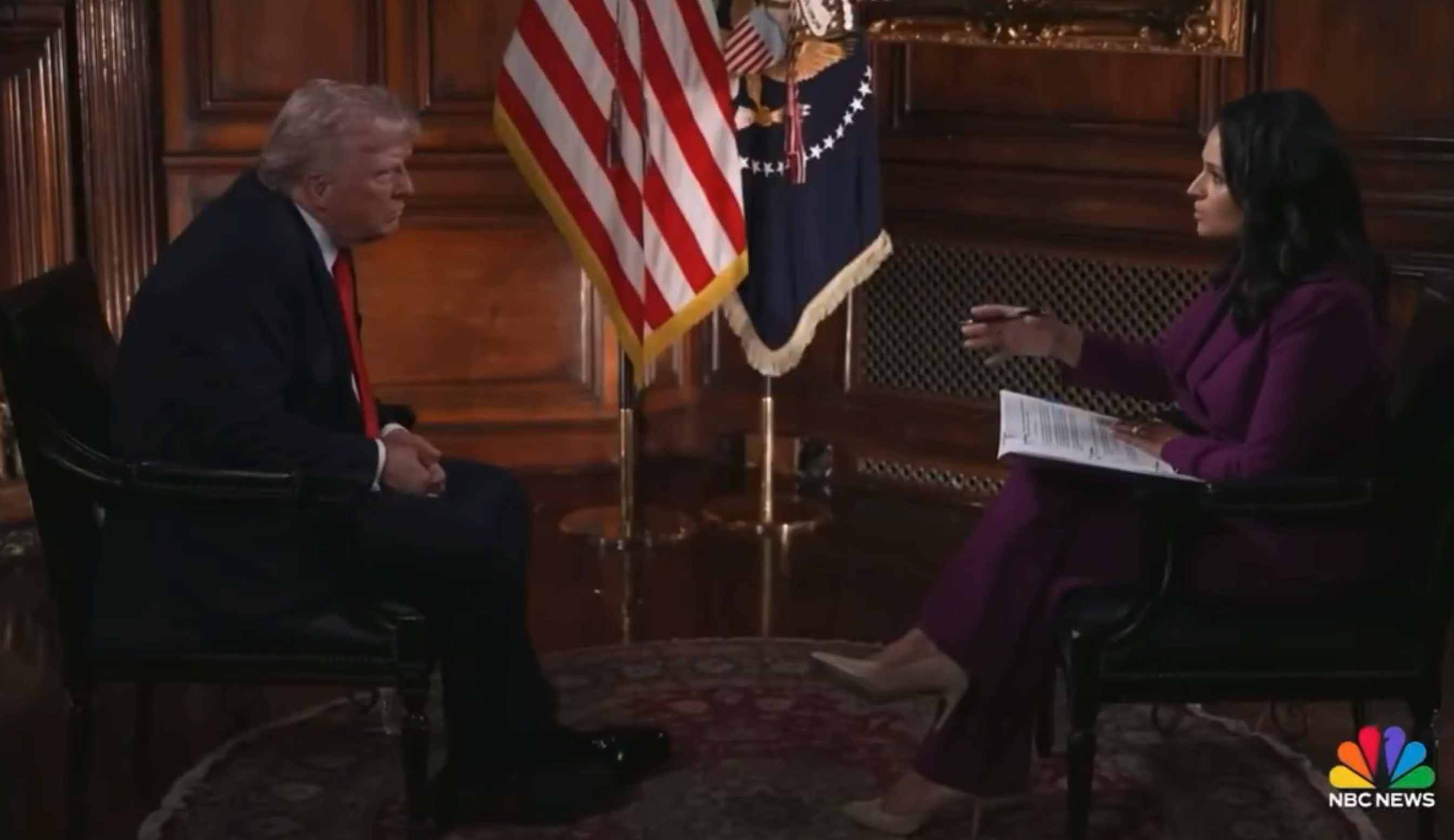 A man and woman sit facing each other during a formal interview in a room with U.S. flags. The woman holds a notepad and pen
