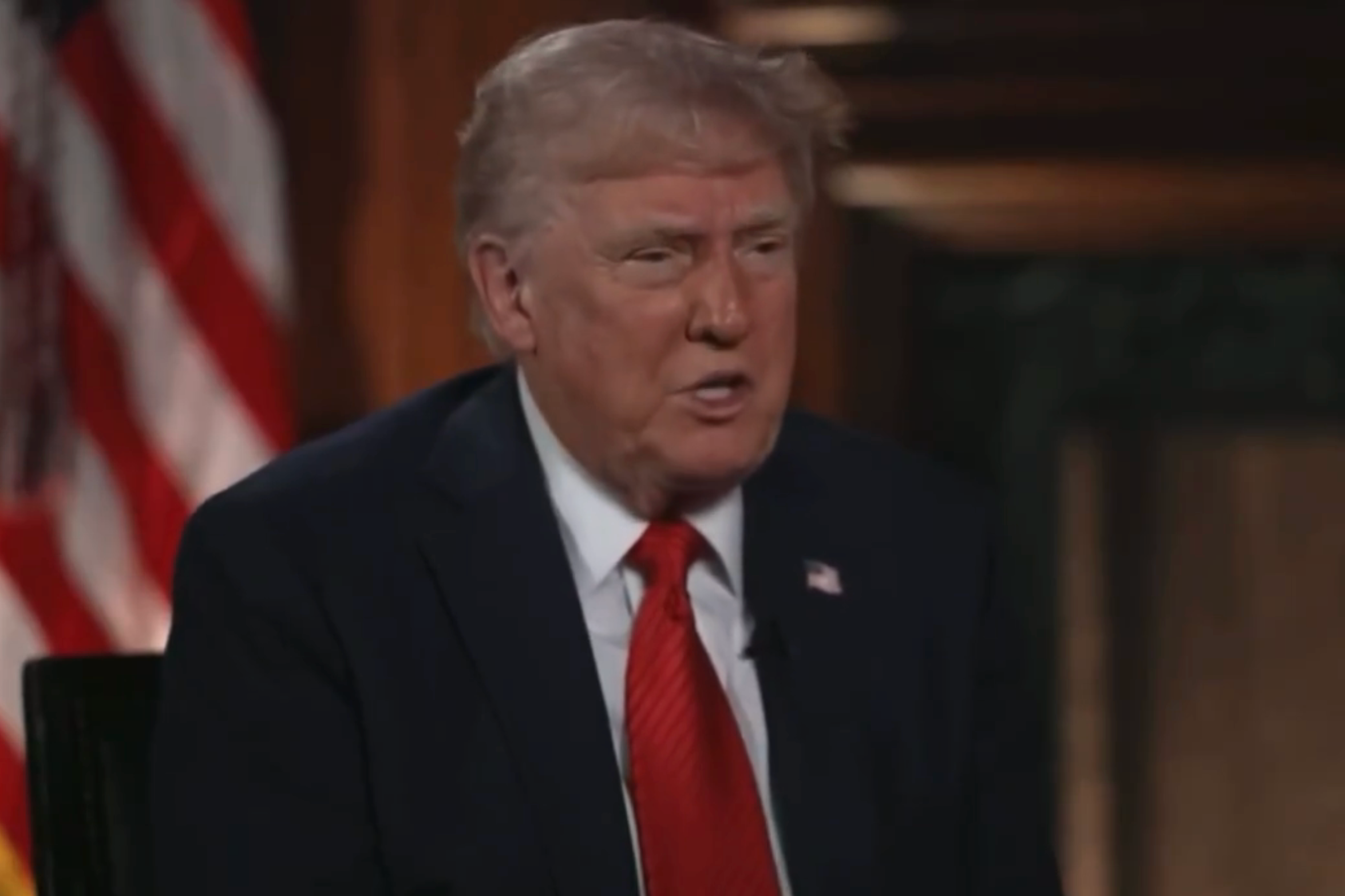 A person in a suit and tie sits for an interview with an American flag in the background