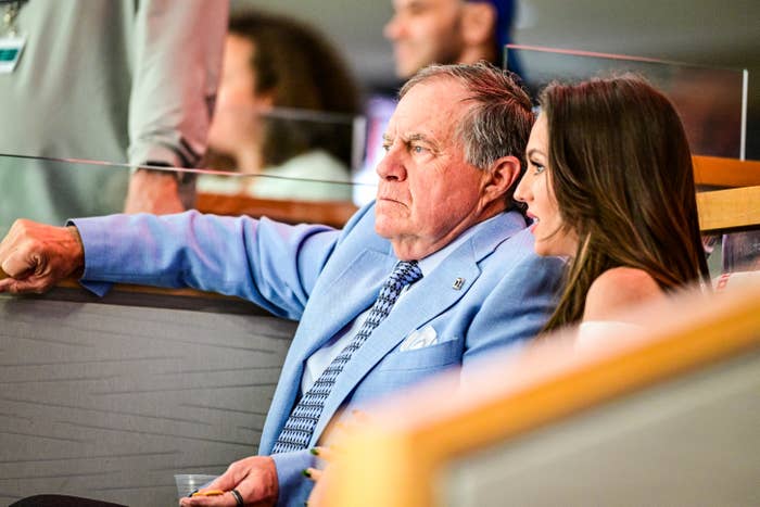 Bill Belichick in a blue suit and Jordon Hudson are seated in a stadium box, engaged in conversation, possibly watching an event