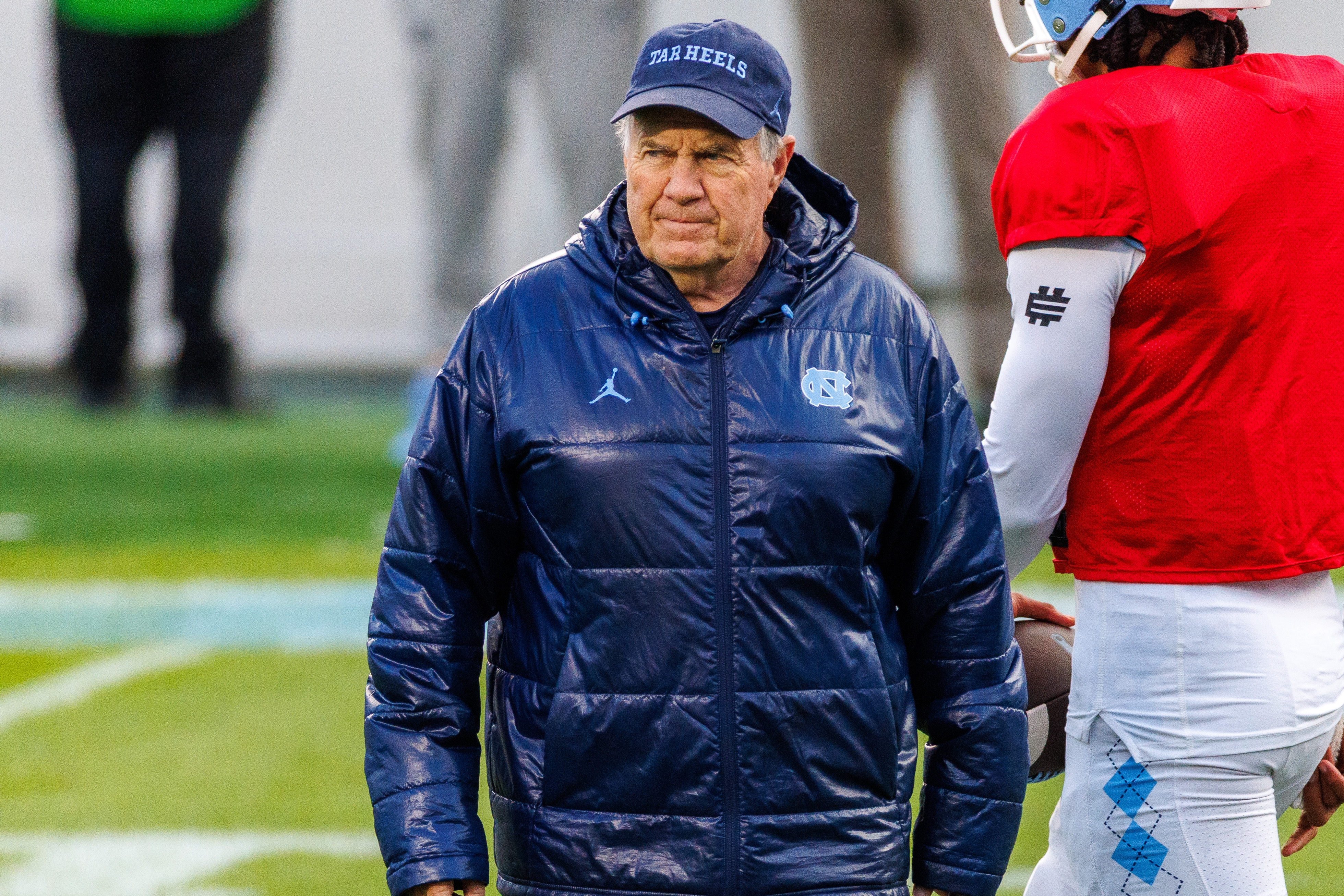 Bill Belichick in a hooded jacket and cap walks on a football field during practice, with players in gear nearby
