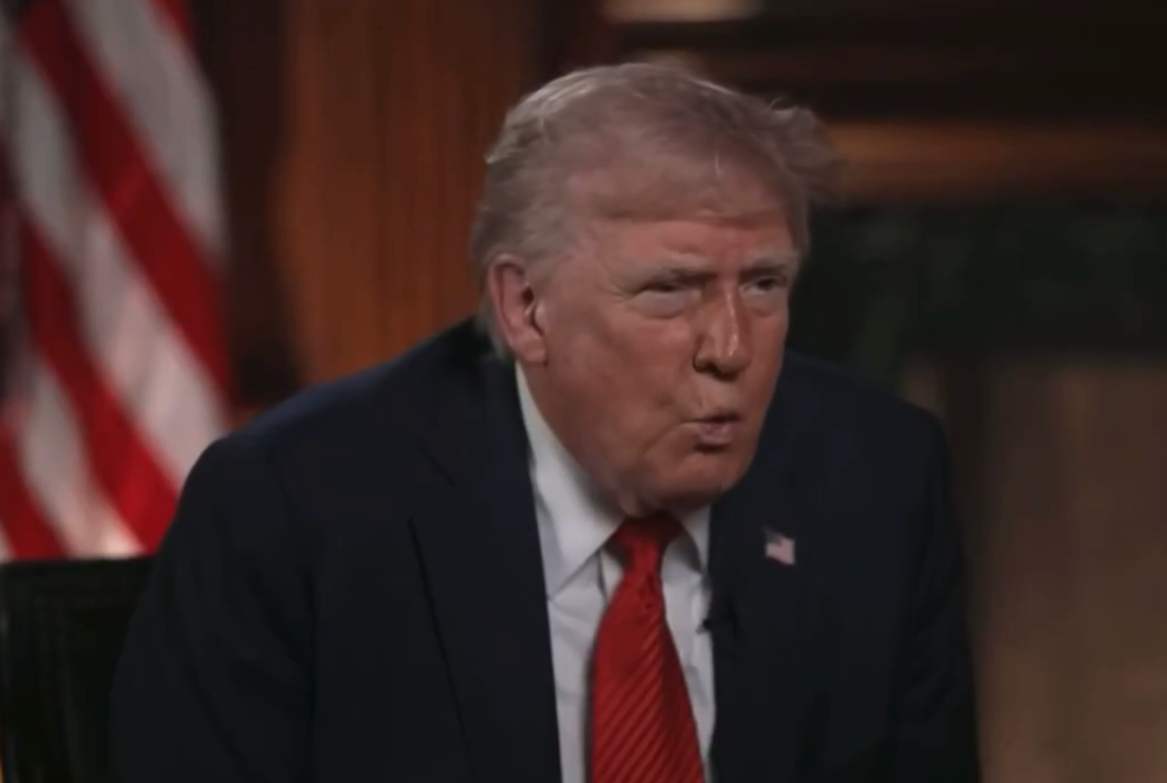 A person in a suit is seated indoors, appearing to speak during an interview. An American flag is visible in the background