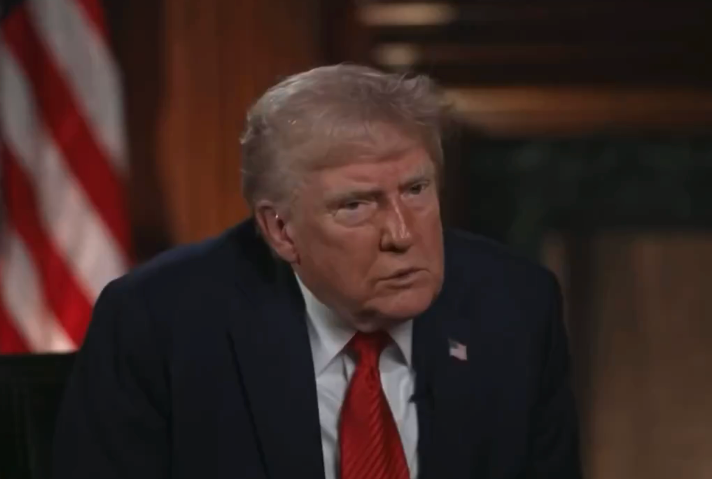 A person in a suit and red tie is seated indoors, appearing focused, with a U.S. flag in the background