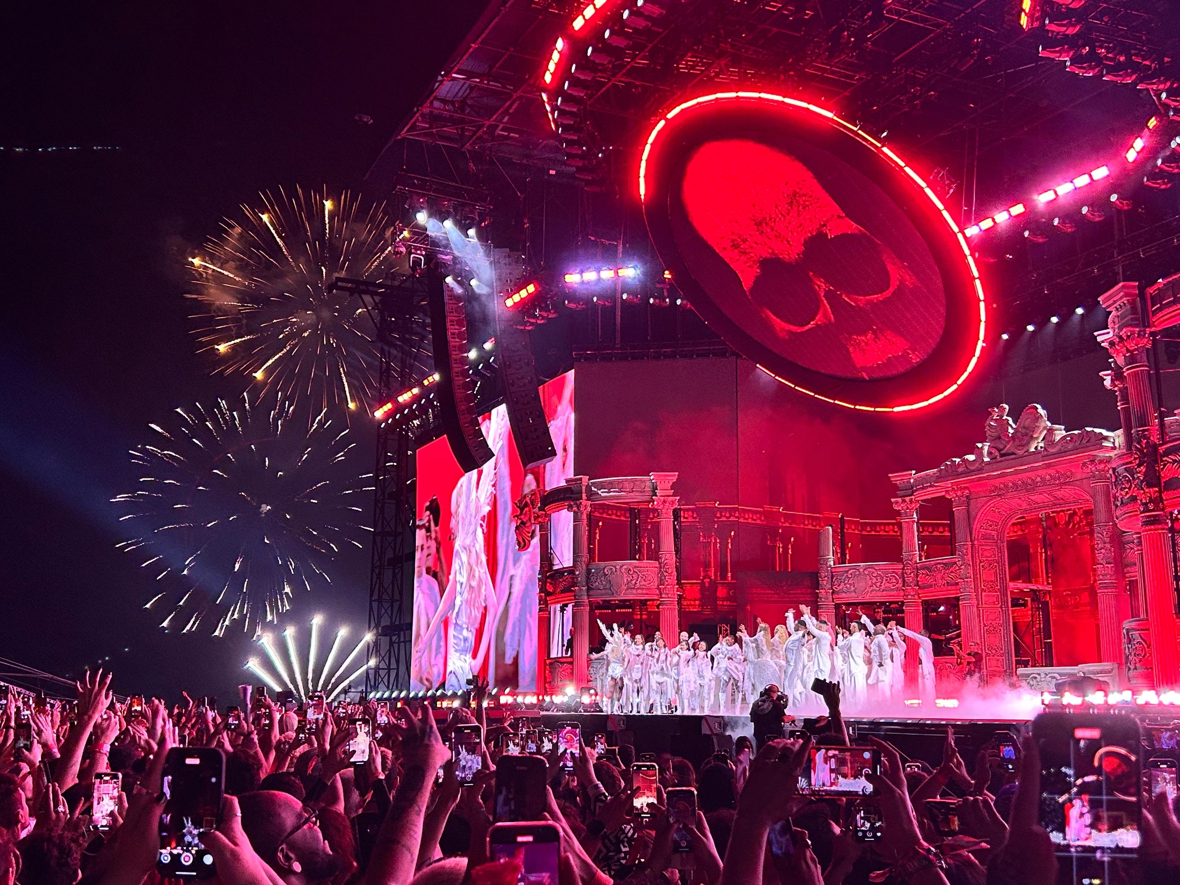 Concert scene with fireworks and a large skull image on screen. Performers in white outfits stand on stage amid crowd taking photos