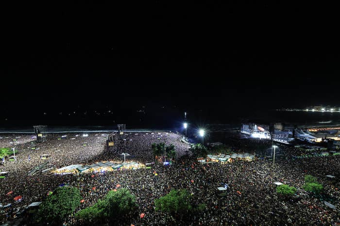 Large crowd gathers for an outdoor nighttime concert by the beach, with lights illuminating the stage and audience