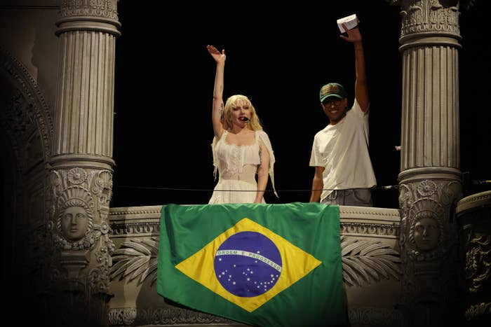 Lady Gaga in elegant white dress and another in casual outfit wave from a balcony adorned with a Brazilian flag