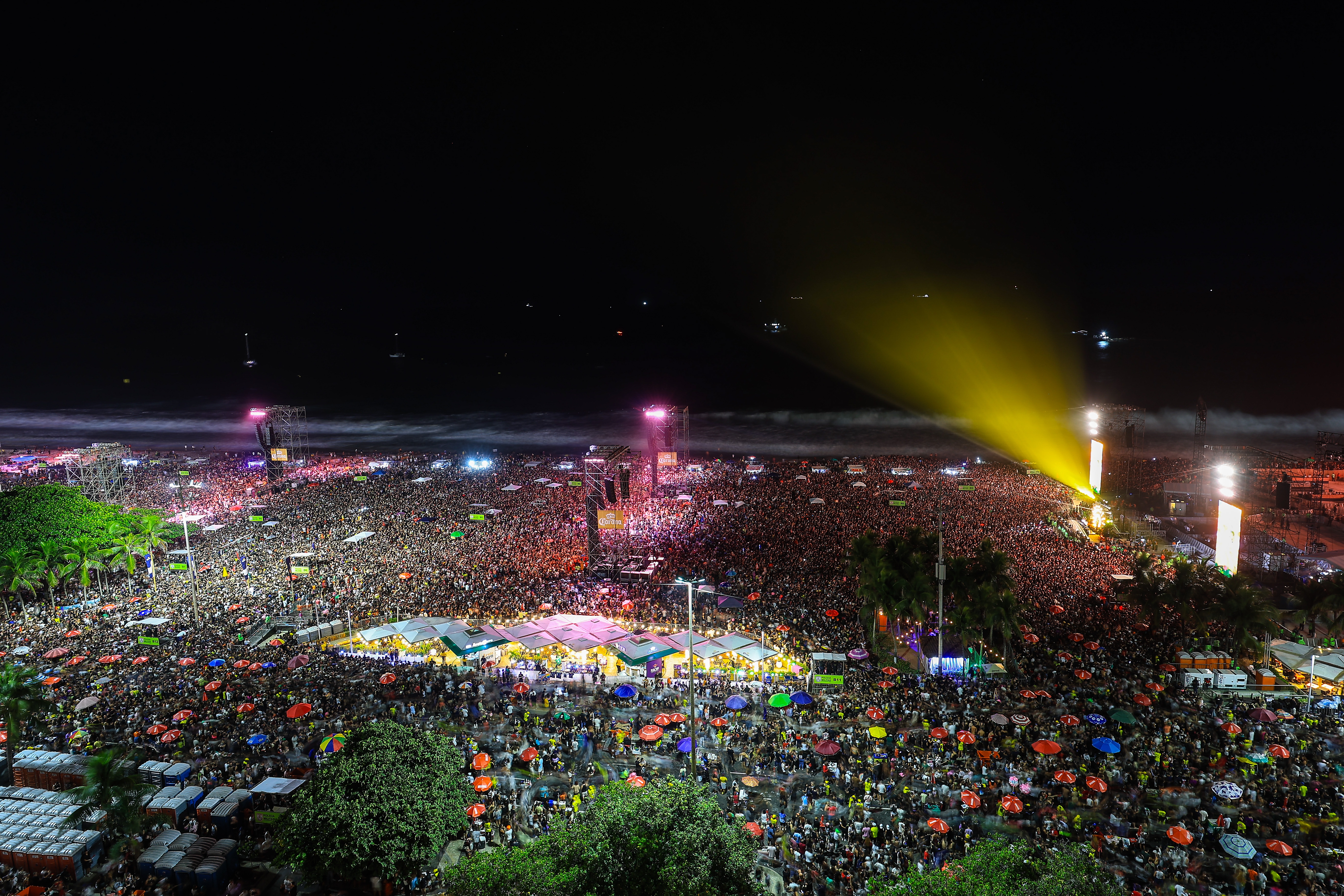 A large, dense crowd enjoys a night concert on a beach, with bright lights and vibrant energy