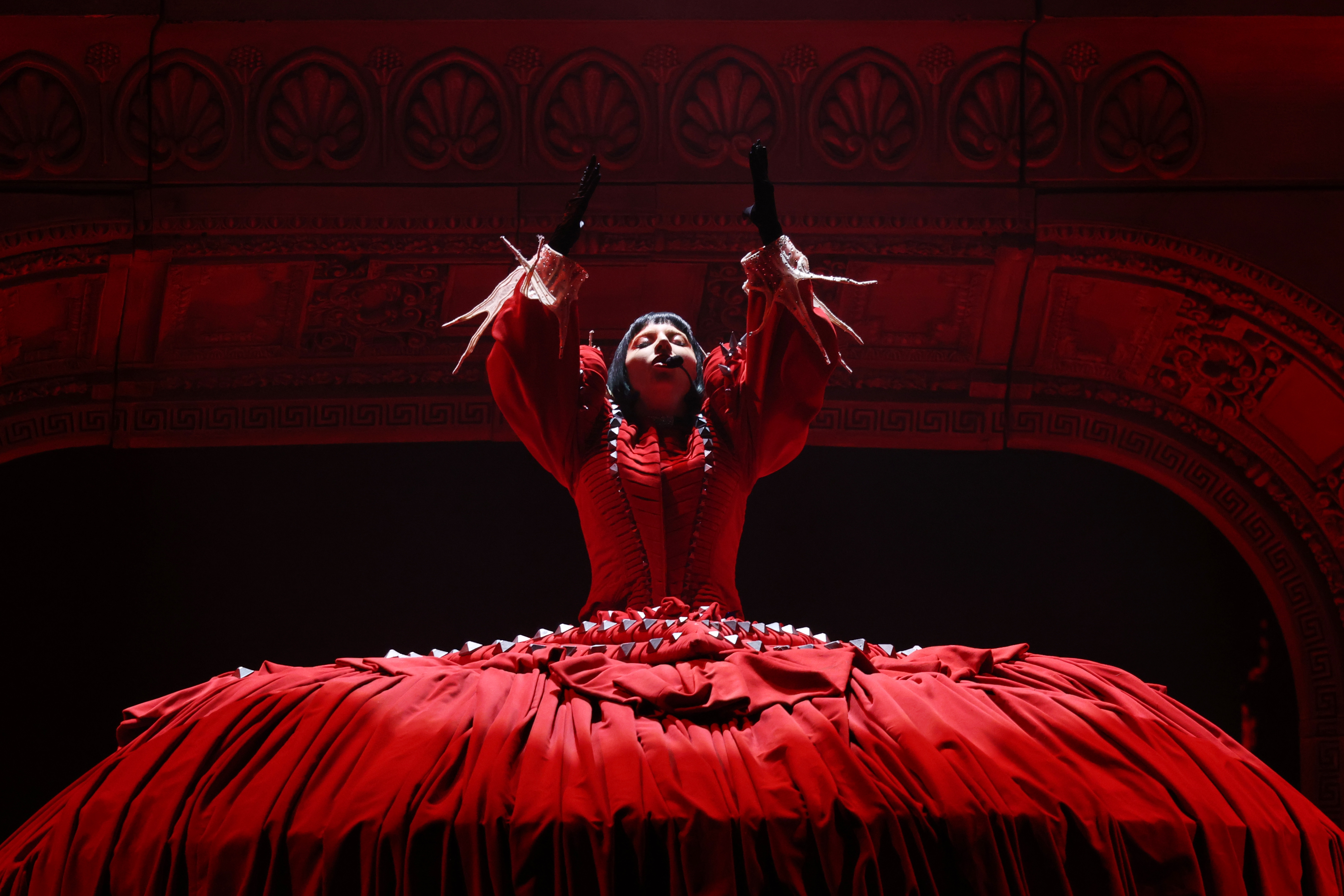 Lady Gaga on stage in an elaborate, voluminous gown with dramatic sleeves, raising arms under ornate ceiling, creating a theatrical atmosphere