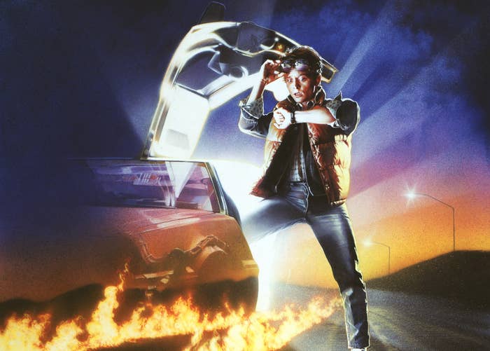 Person in a vest stands beside a car with gullwing doors open, looking at a watch. Flames trail behind the car on a road