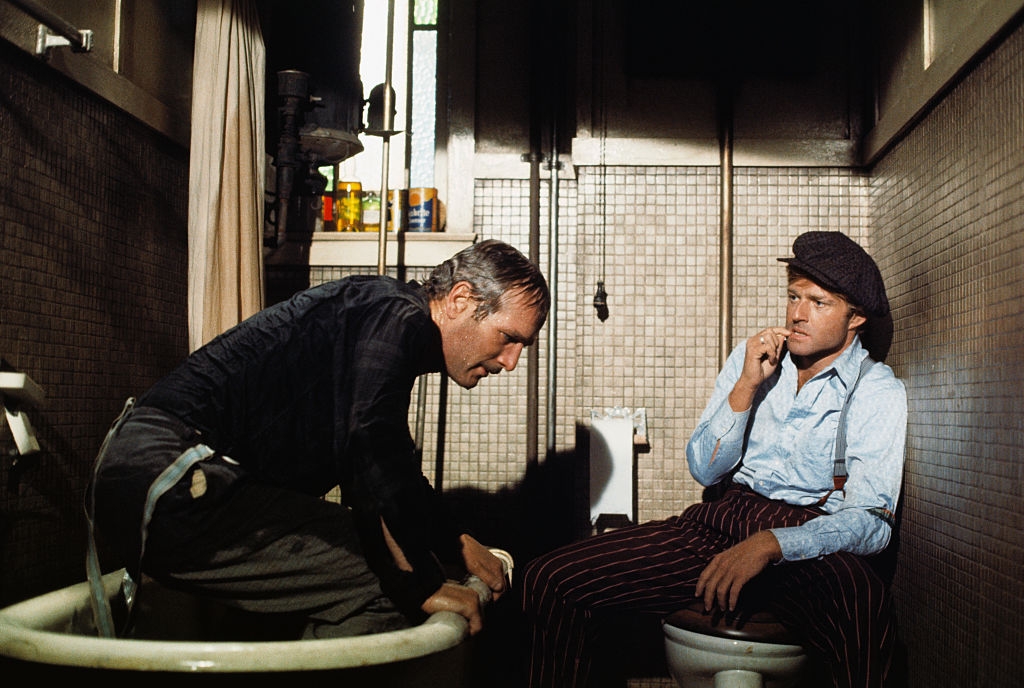 Two men in an old-fashioned bathroom, one sits on the toilet in a cap and striped pants; the other leans over a bathtub, appearing engaged in conversation