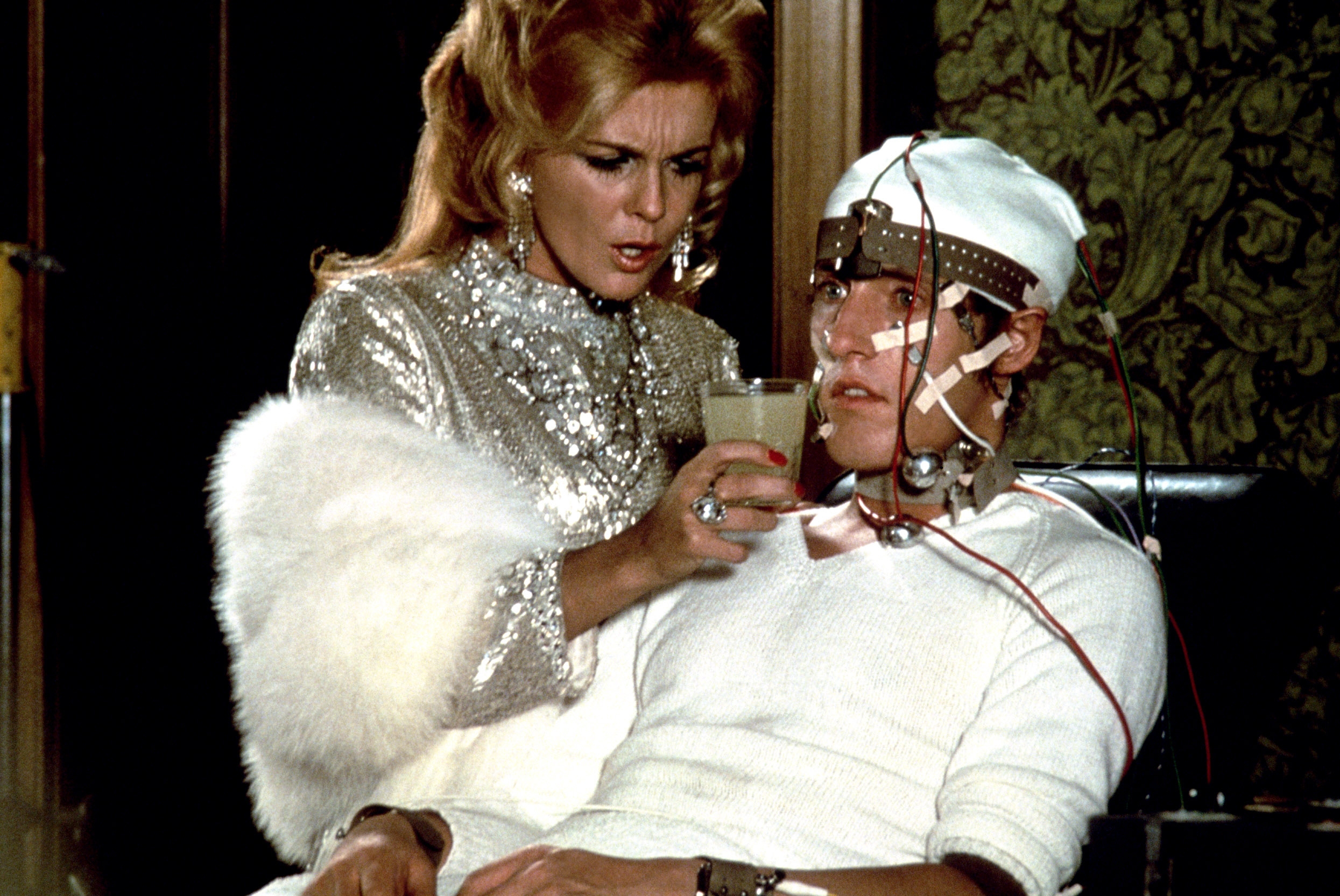 A woman in glamorous attire offers a drink to a man in a mechanical restraint chair with wires attached, depicting a scene from a sci-fi movie