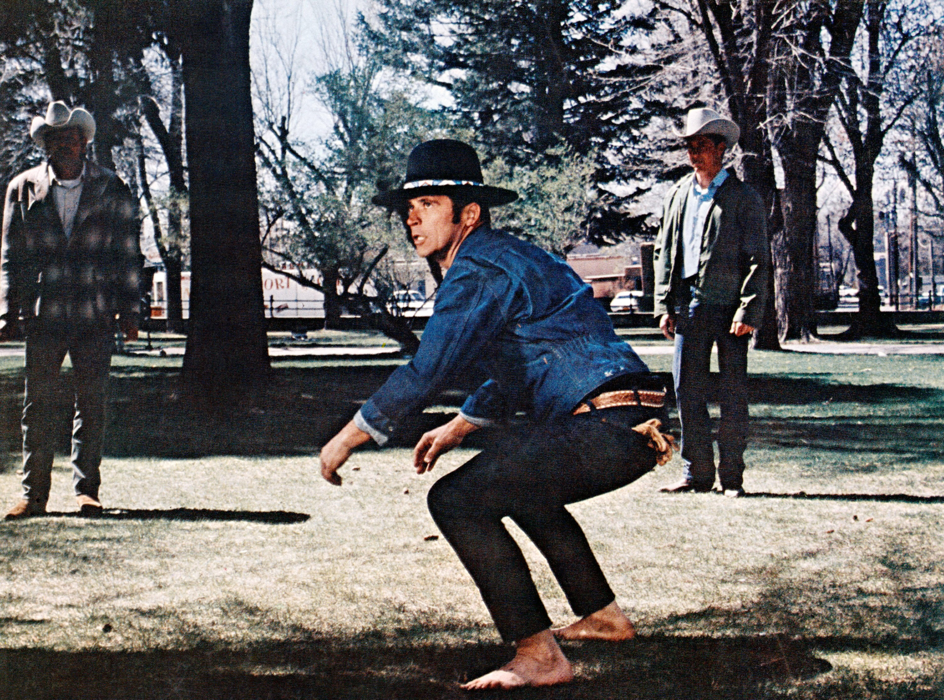 A person in a denim outfit squats barefoot on grass in a park, with two others in cowboy hats observing. Trees and park structures in the background
