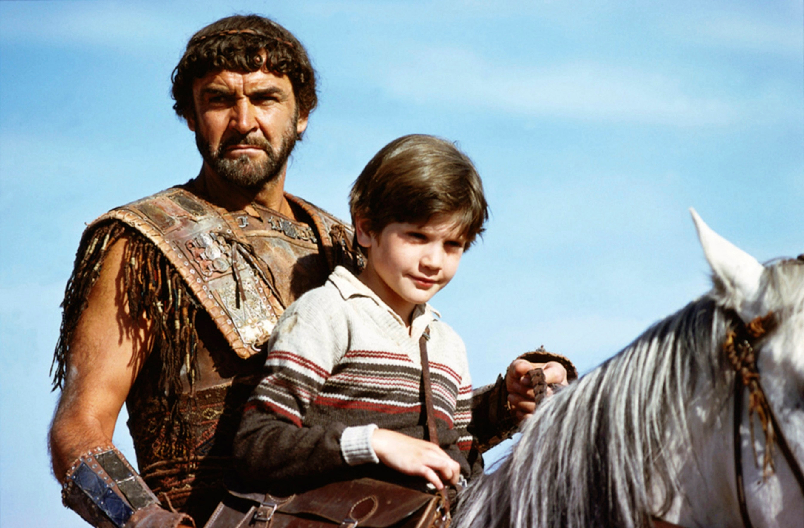 Man in rugged, ancient-style costume with young boy, both on horseback, in a historical film setting